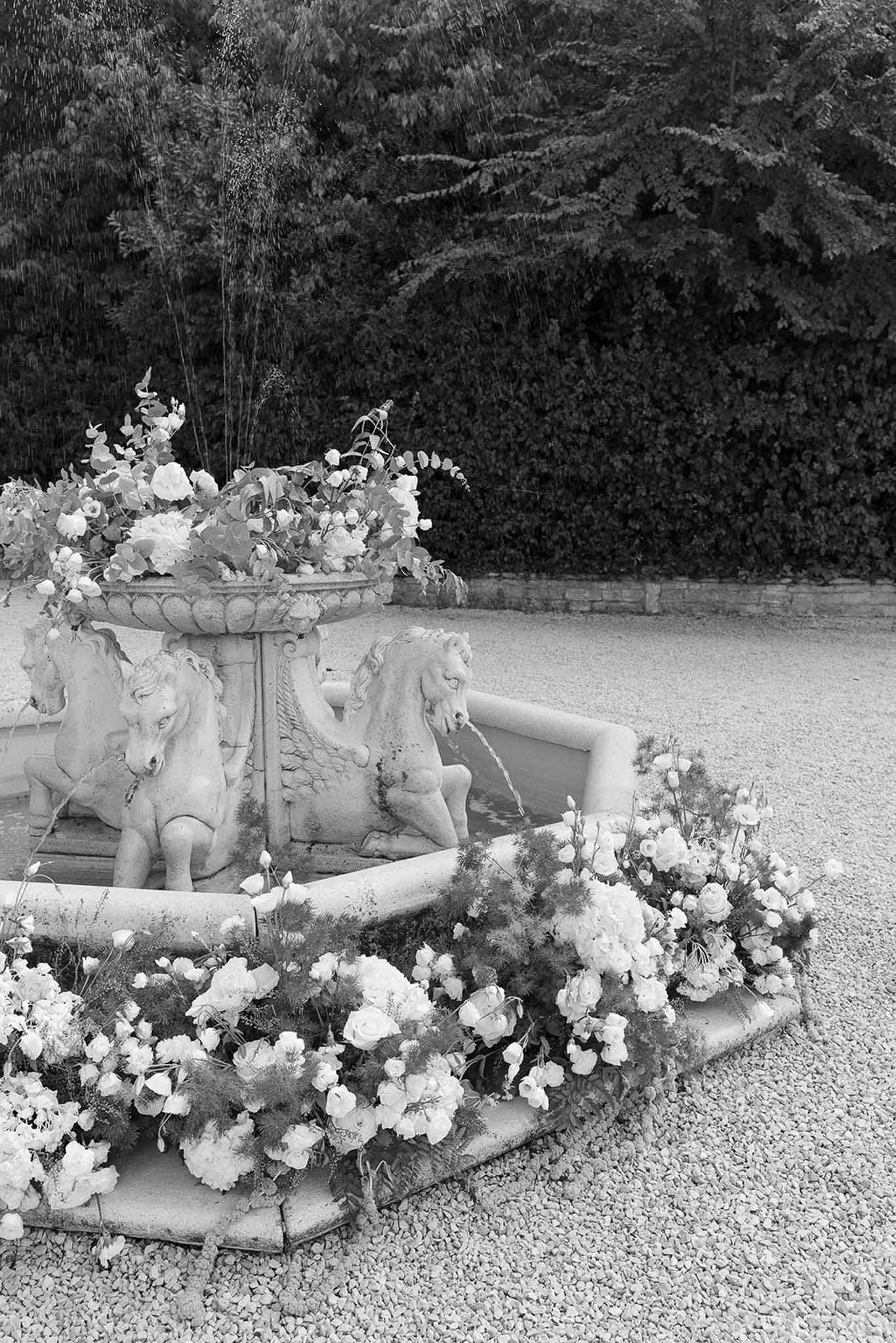 Black-and-white image of ornate stone fountain decorated with roses and trailing greenery on a chateau courtyard