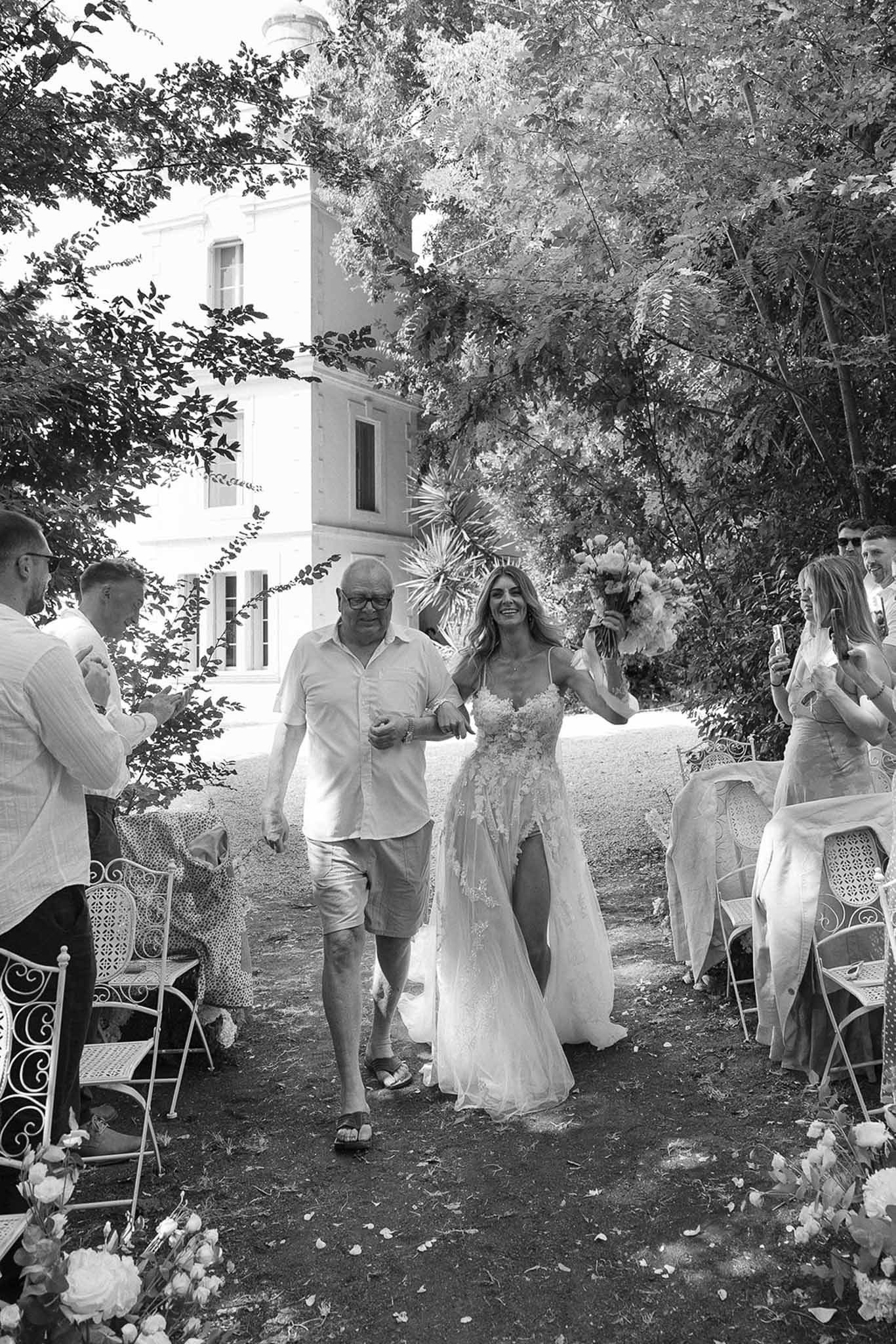 Black-and-white image of bride walking down garden aisle with father as guests watch at French chateau ceremony