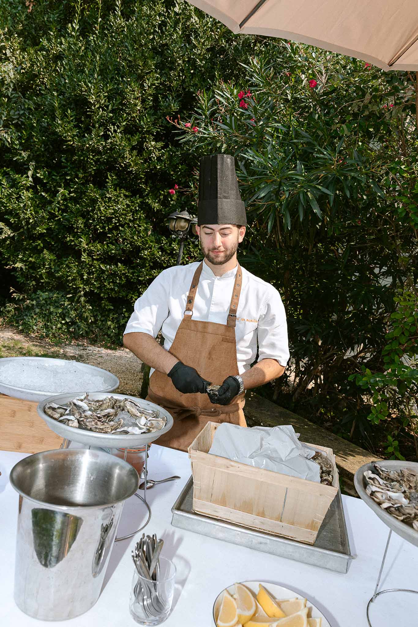 Chef in white jacket shucking oysters at outdoor cocktail station with tiered seafood stands and lemon wedges