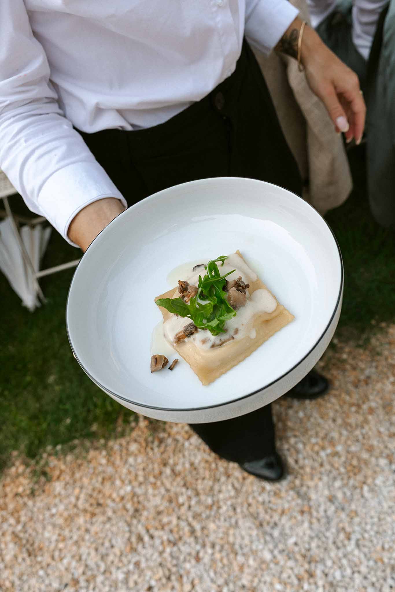 Server carrying plated ravioli with cream sauce mushrooms and arugula at outdoor reception