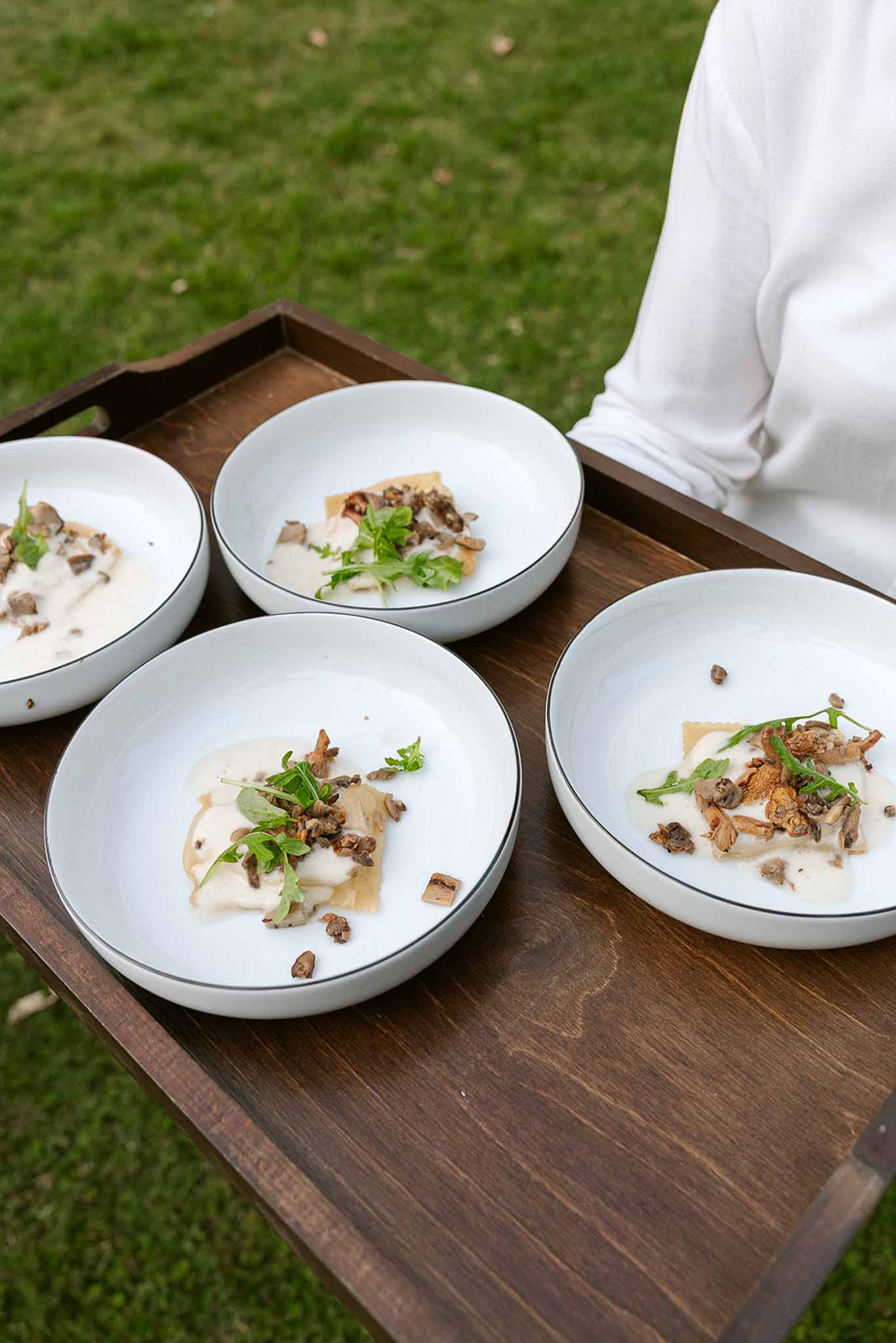 Four white bowls of ravioli with wild mushrooms and arugula on dark wood serving tray held by server