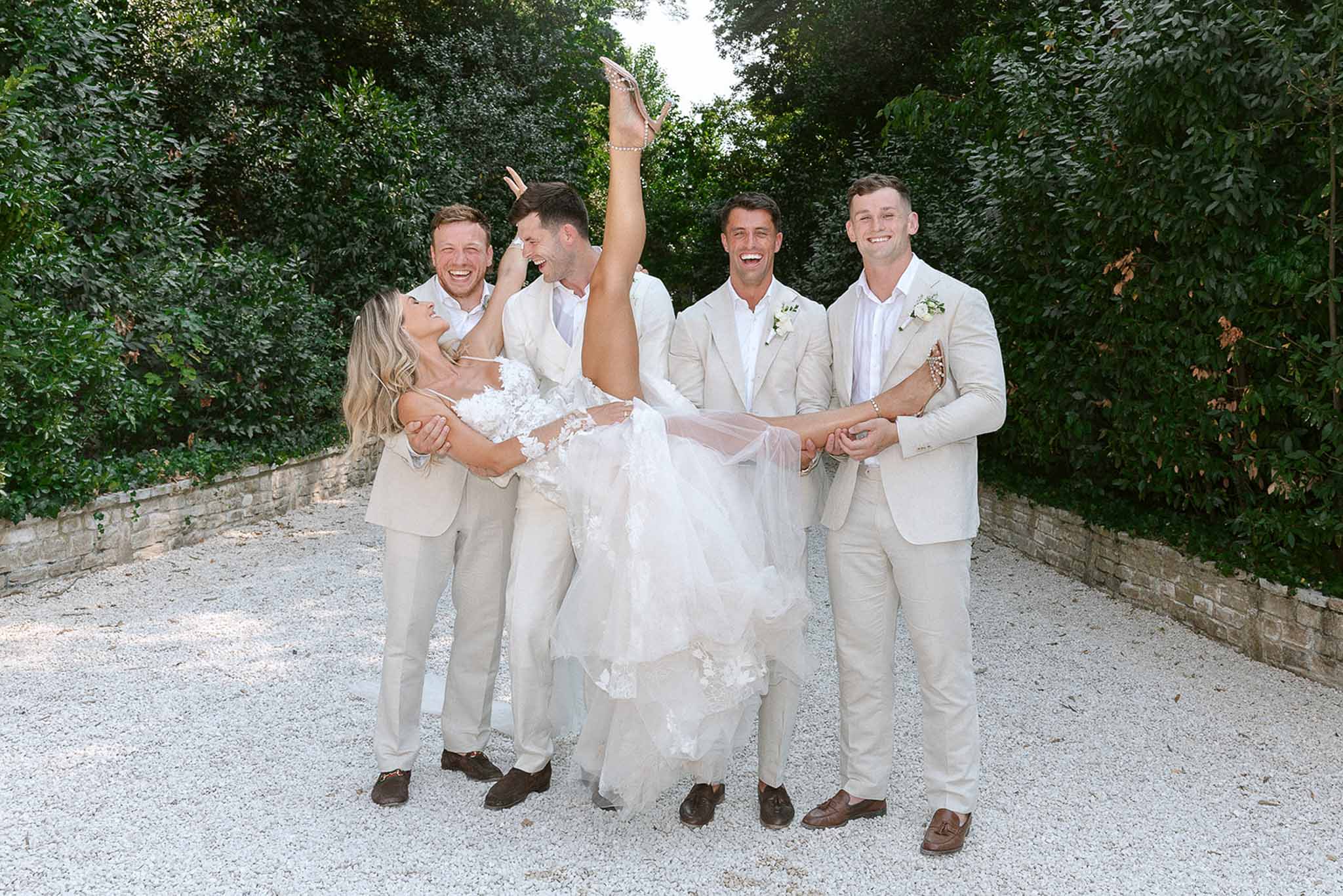 Four groomsmen in cream linen suits lifting bride horizontally on gravel path with hedging