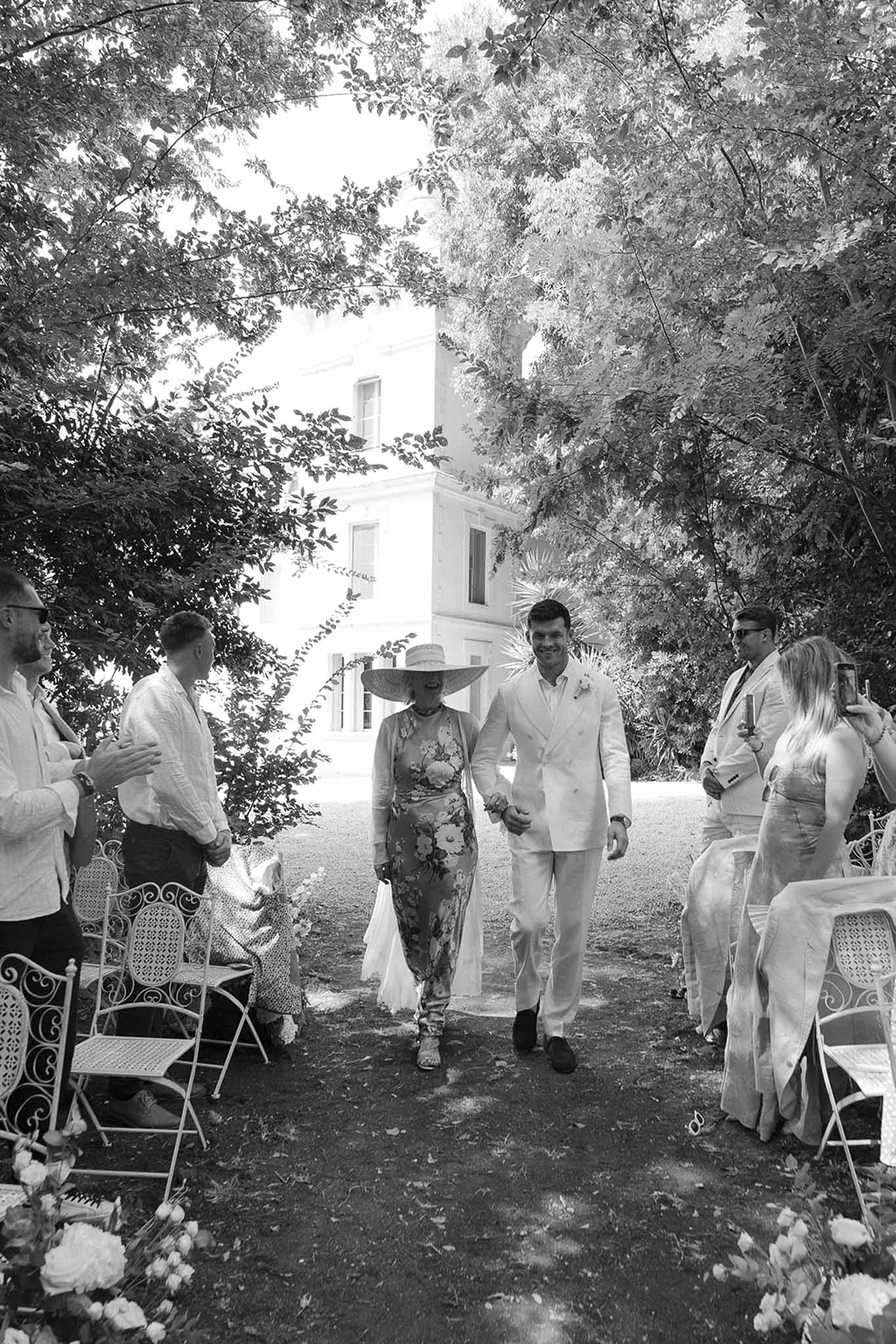 Black and white processional of groom escorted by woman in hat down grass aisle at chateau grounds