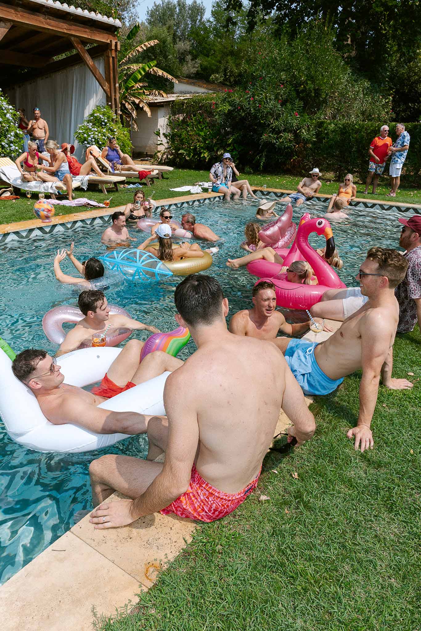 Thirty guests with colourful pool floats at rectangular pool party with pergola and tropical plantings