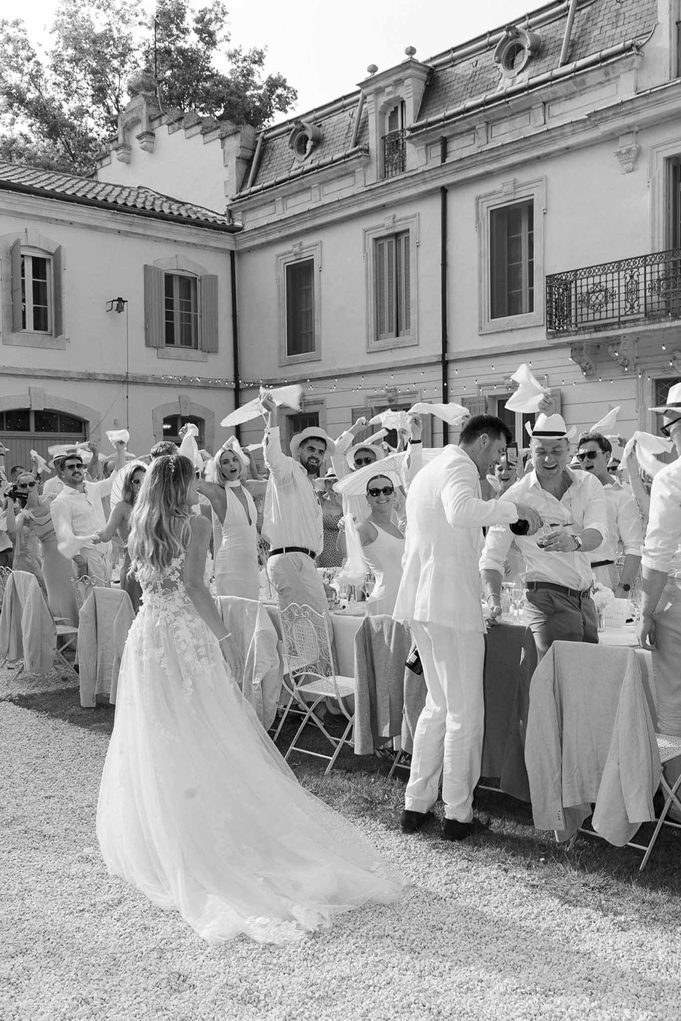 Couple enters between tables as 30 guests wave napkins and white hats in French tradition in B&W
