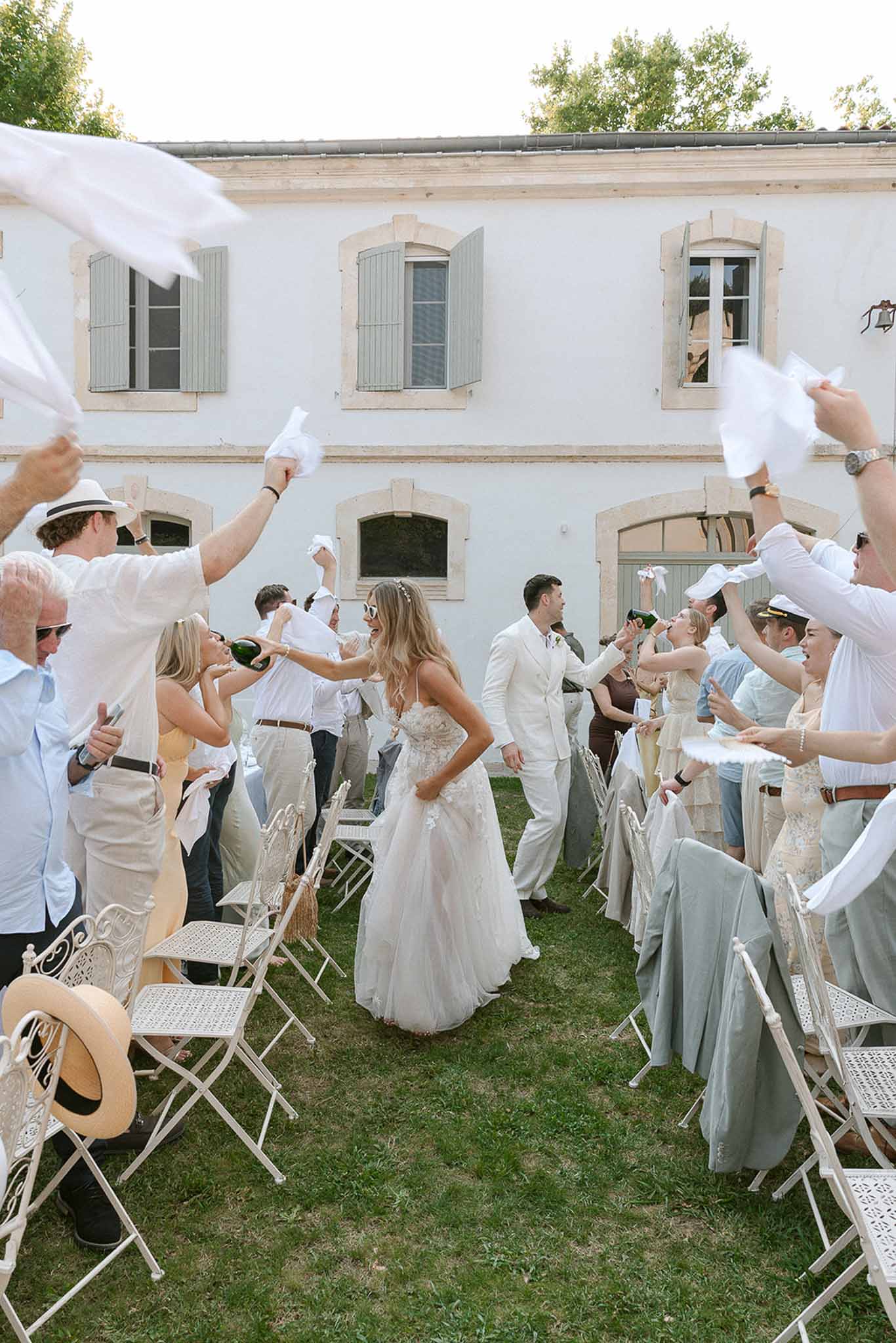 Bride and groom walk through guest tunnel as guests wave white napkins on lawn of French manor house