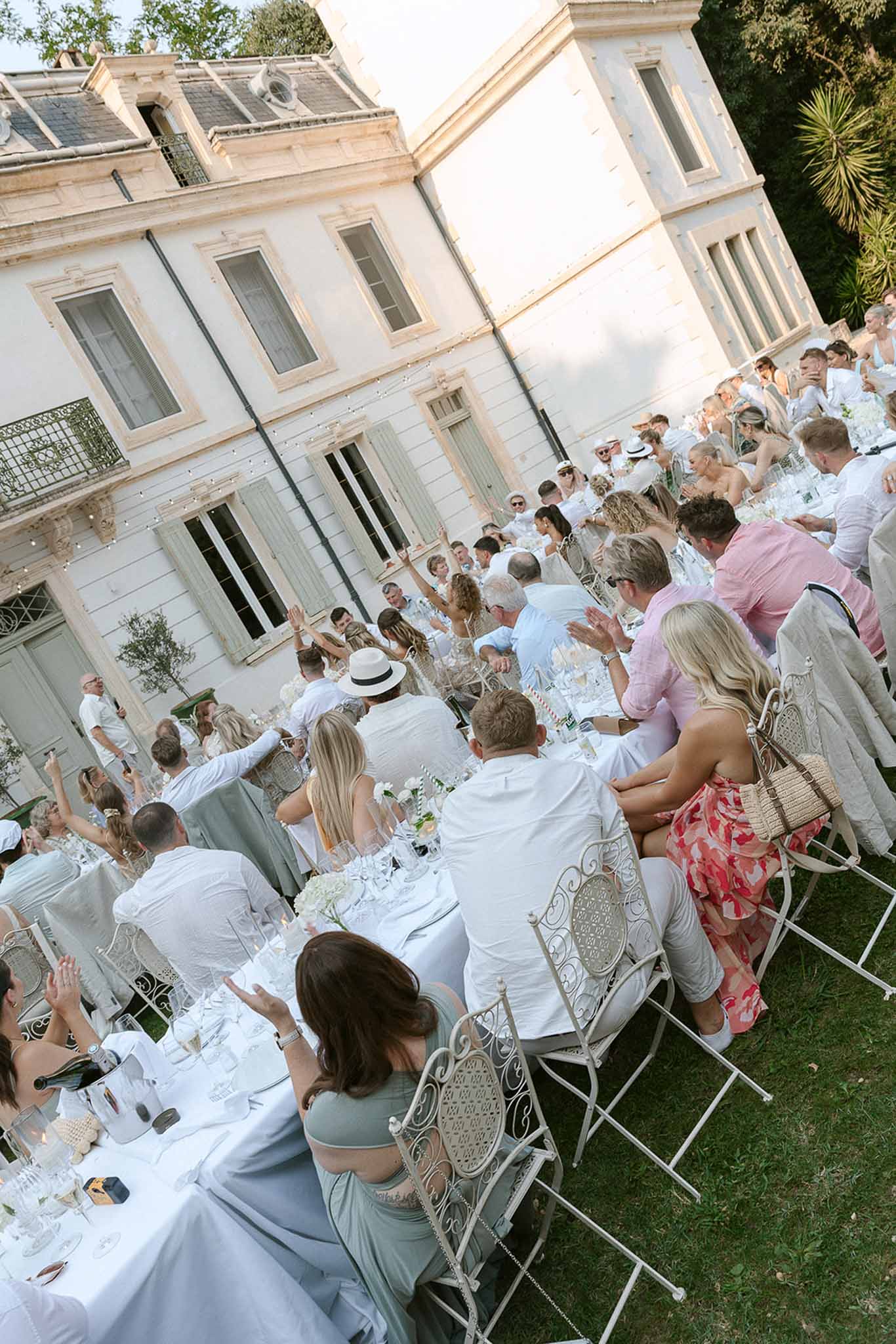 Guests in white dress code clapping at long garden tables with hydrangea centerpieces before villa with fairy lights
