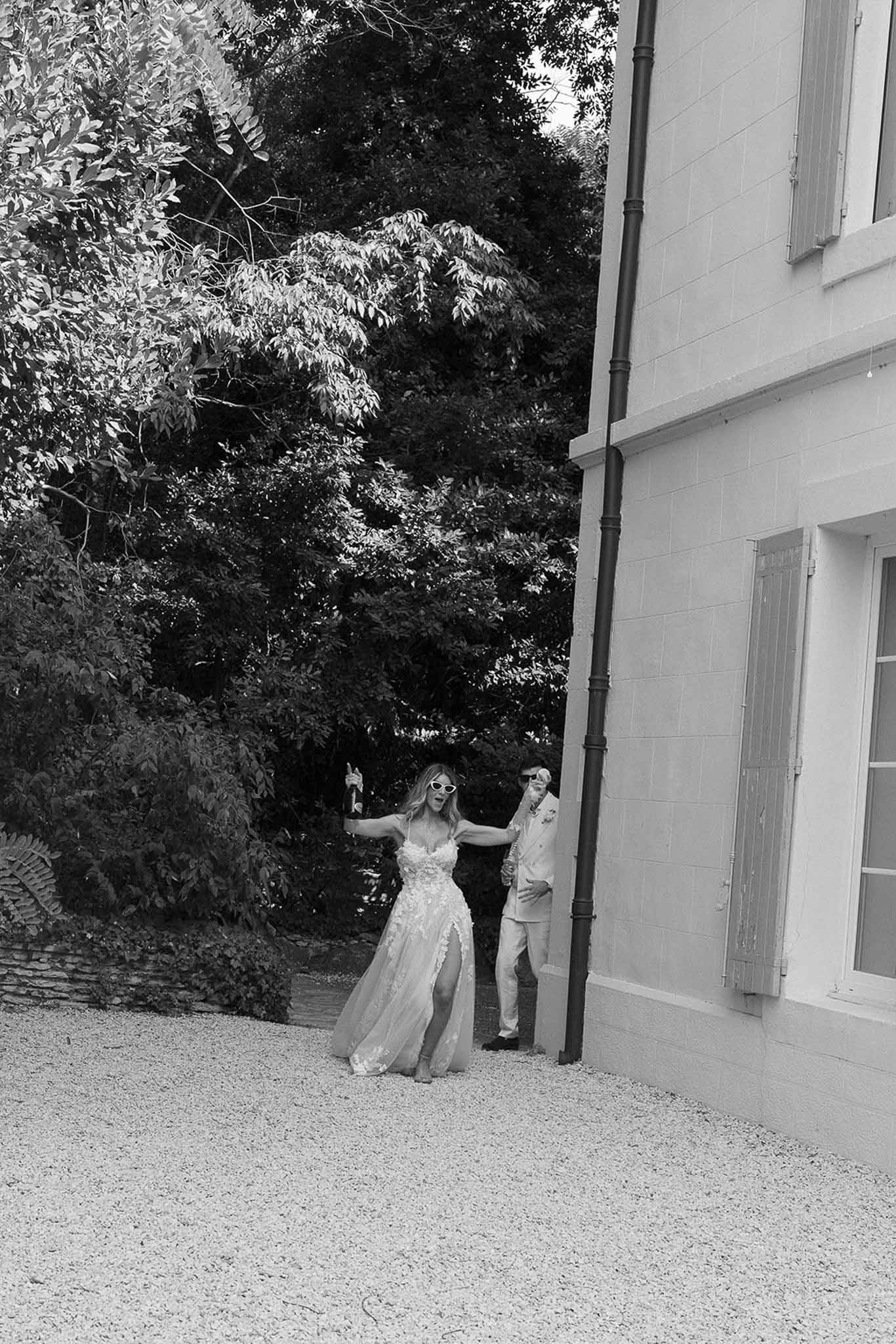 Bride celebrating with champagne bottle on chateau gravel courtyard, groom leaning against building behind