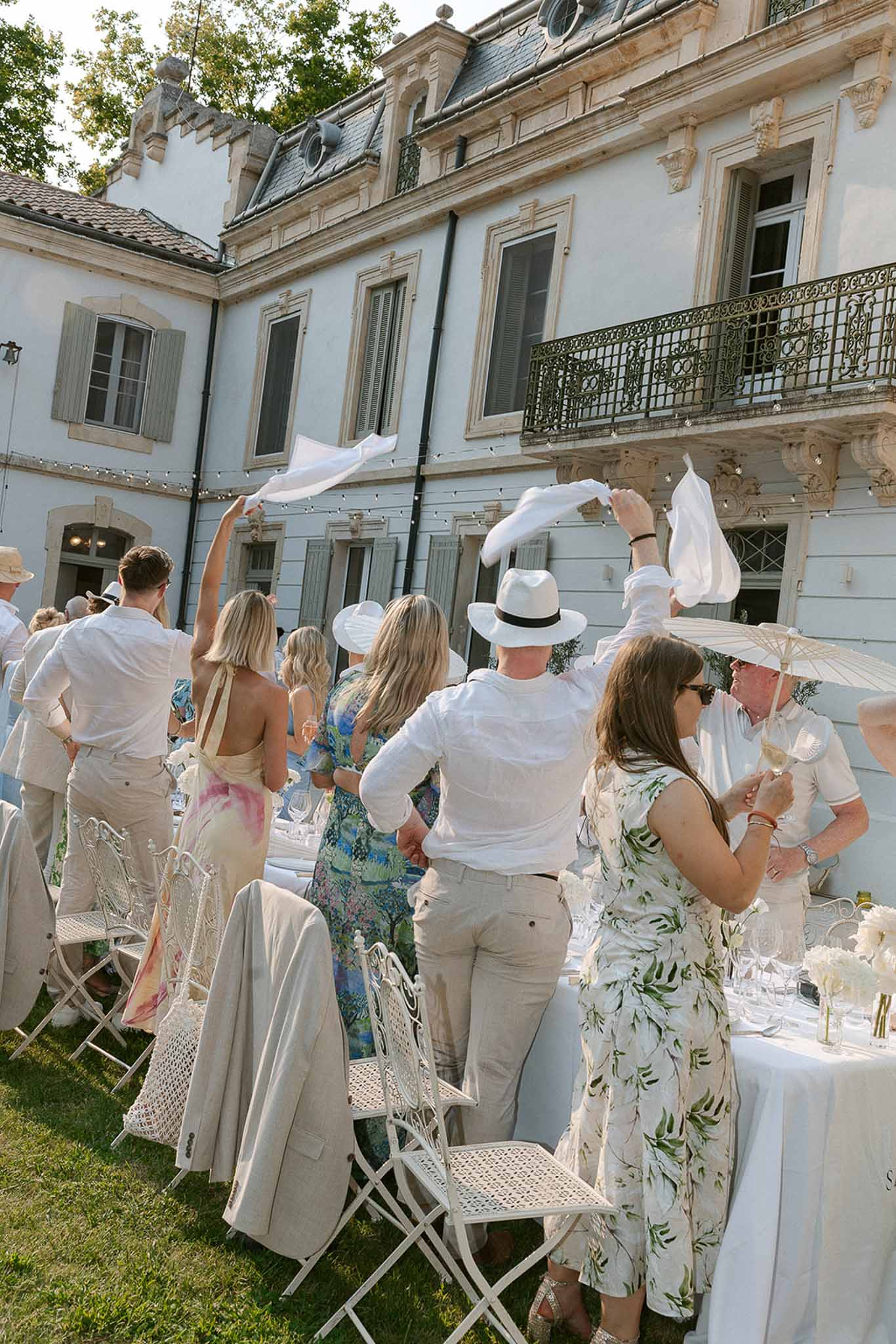 Wedding guests wave white napkins at long banquet tables in chateau courtyard strung with fairy lights