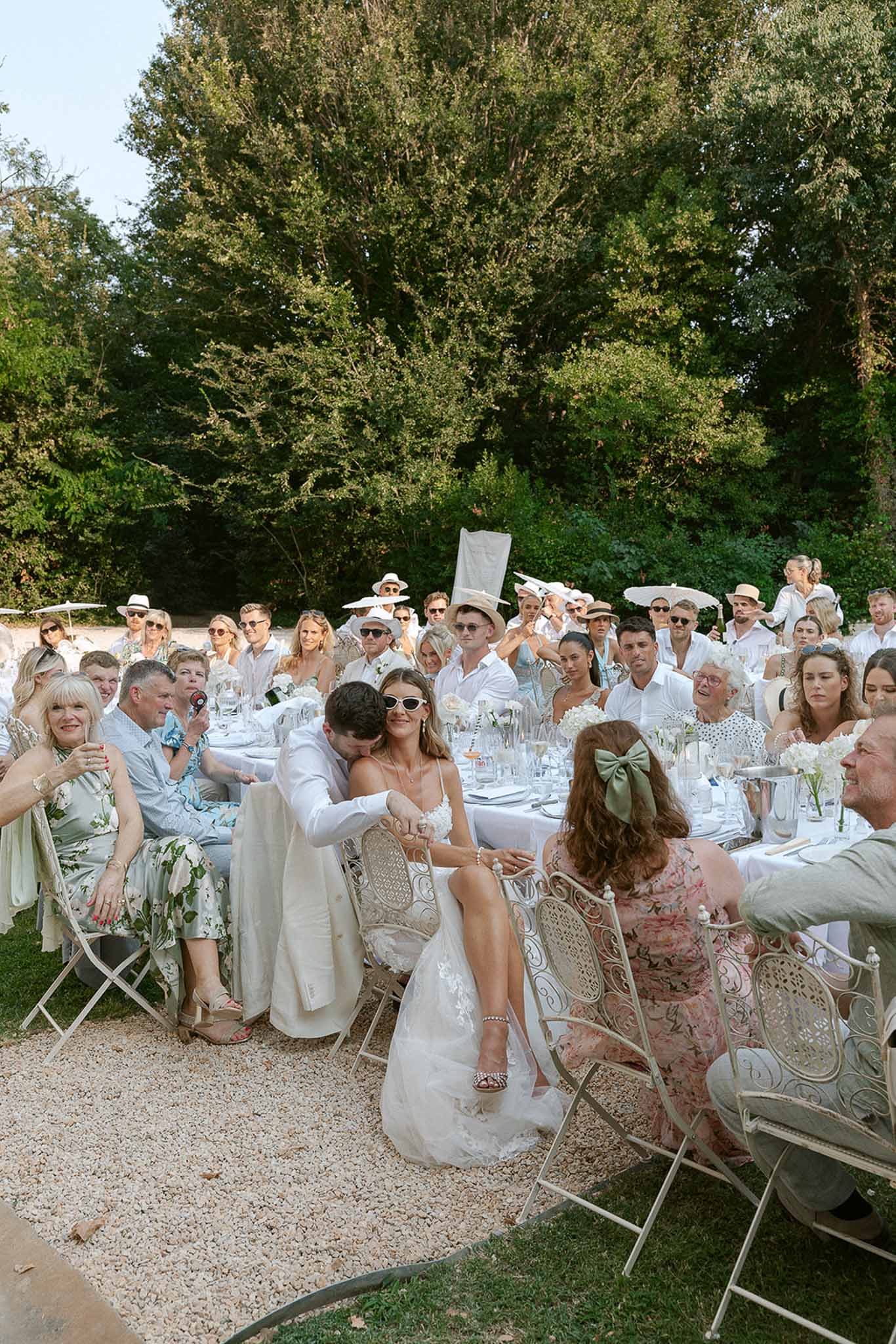 Groom in white suit kisses bride's hand at all-white garden party reception with 45 guests at golden hour