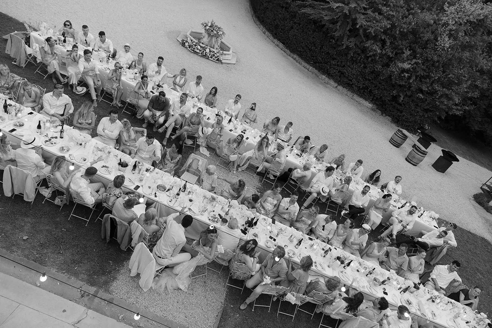 Black-and-white aerial view of outdoor reception with 80-100 guests at long banquet tables on gravel courtyard