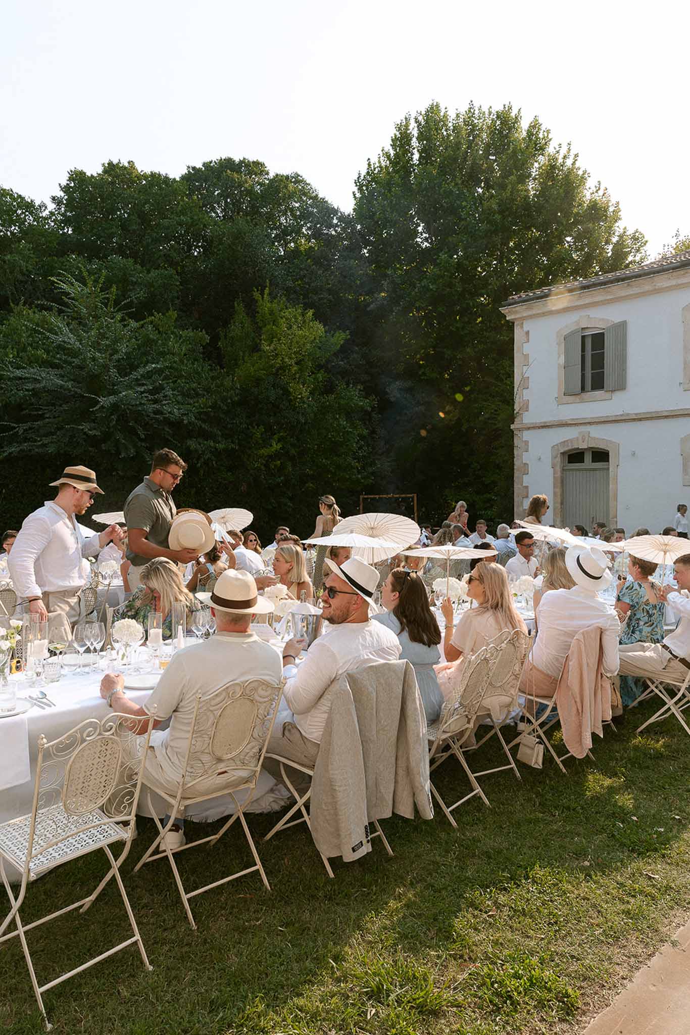 Outdoor reception with guests seated at long white table under parasols beside a French country house