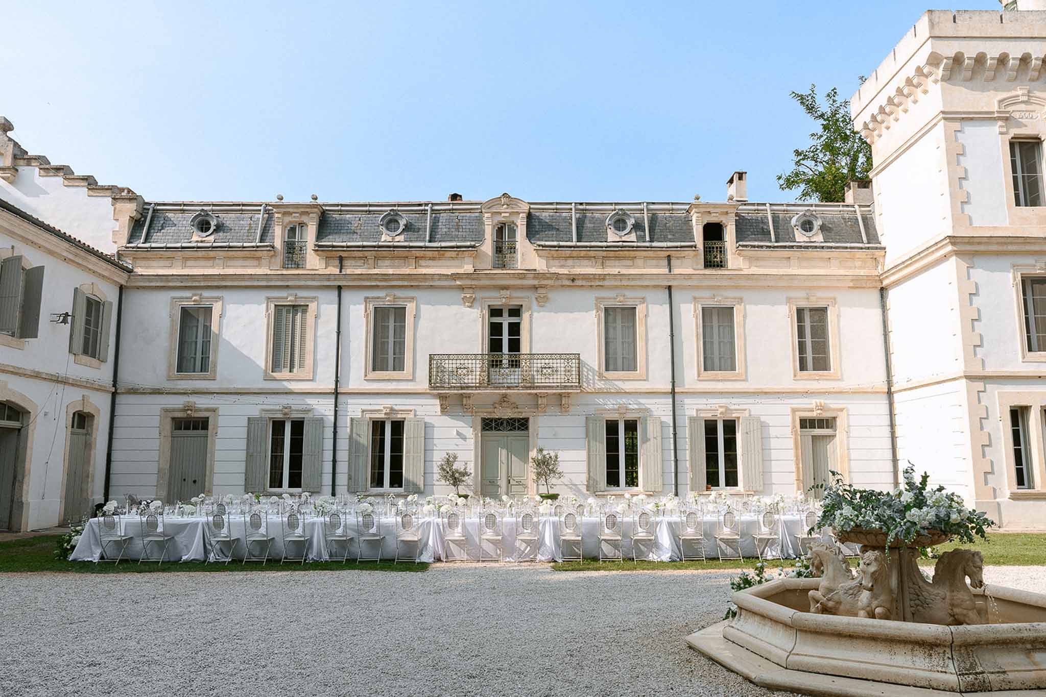 Long tables with ghost chairs and olive trees along chateau facade with white floral horse fountain in foreground