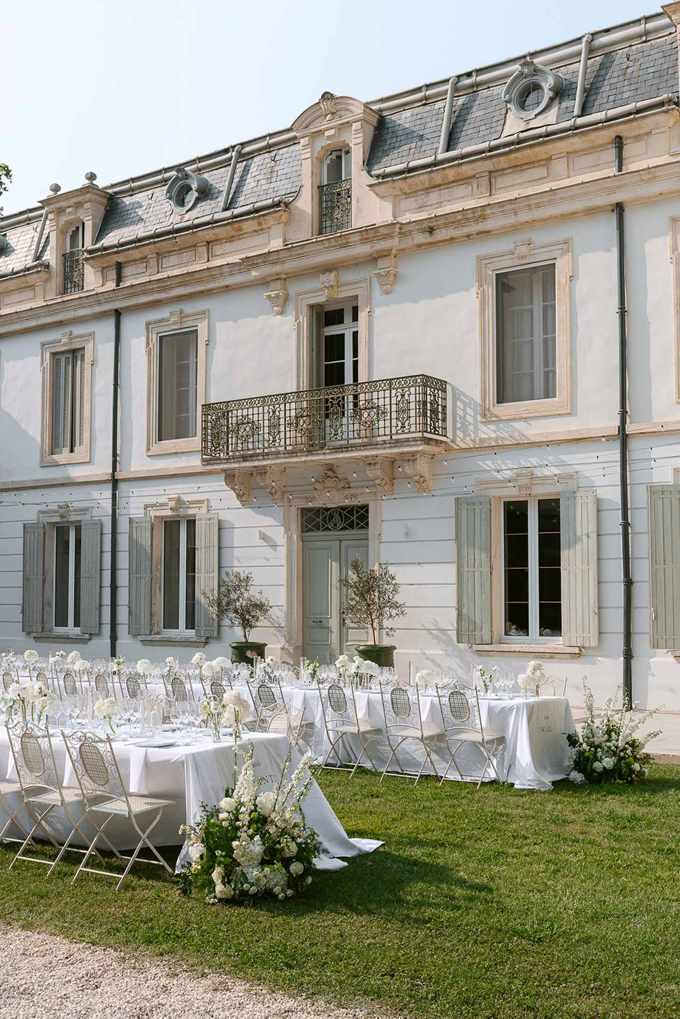 Long white reception tables with rose centrepieces and fairy lights before chateau facade on lawn
