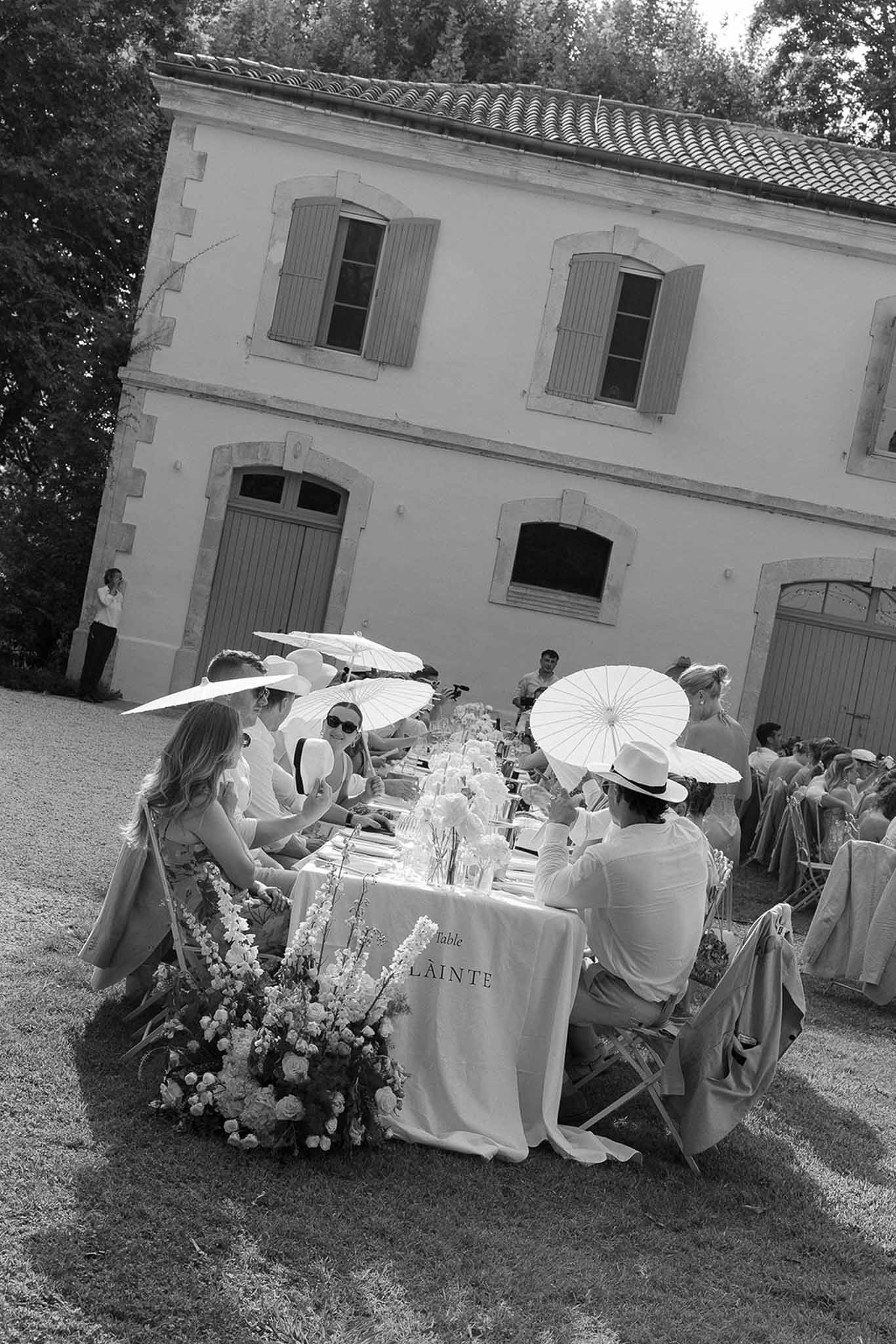 Black-and-white wide shot of guests seated at long banquet table with parasols at French estate reception