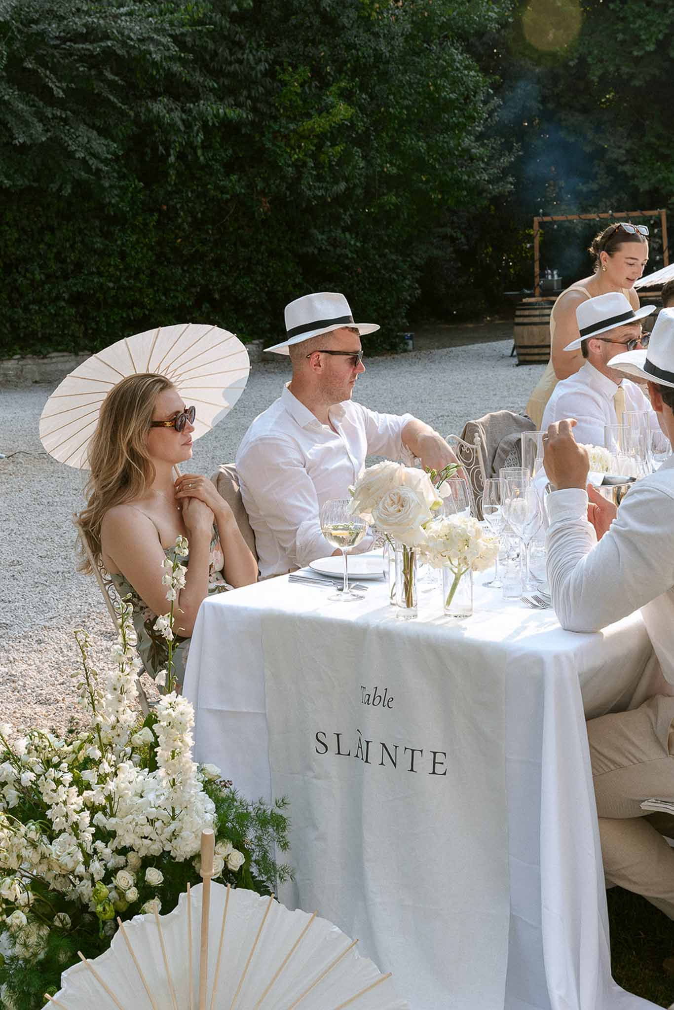 Guests in all-white with Panama hats and parasols at table with ivory roses and sweet pea floor display