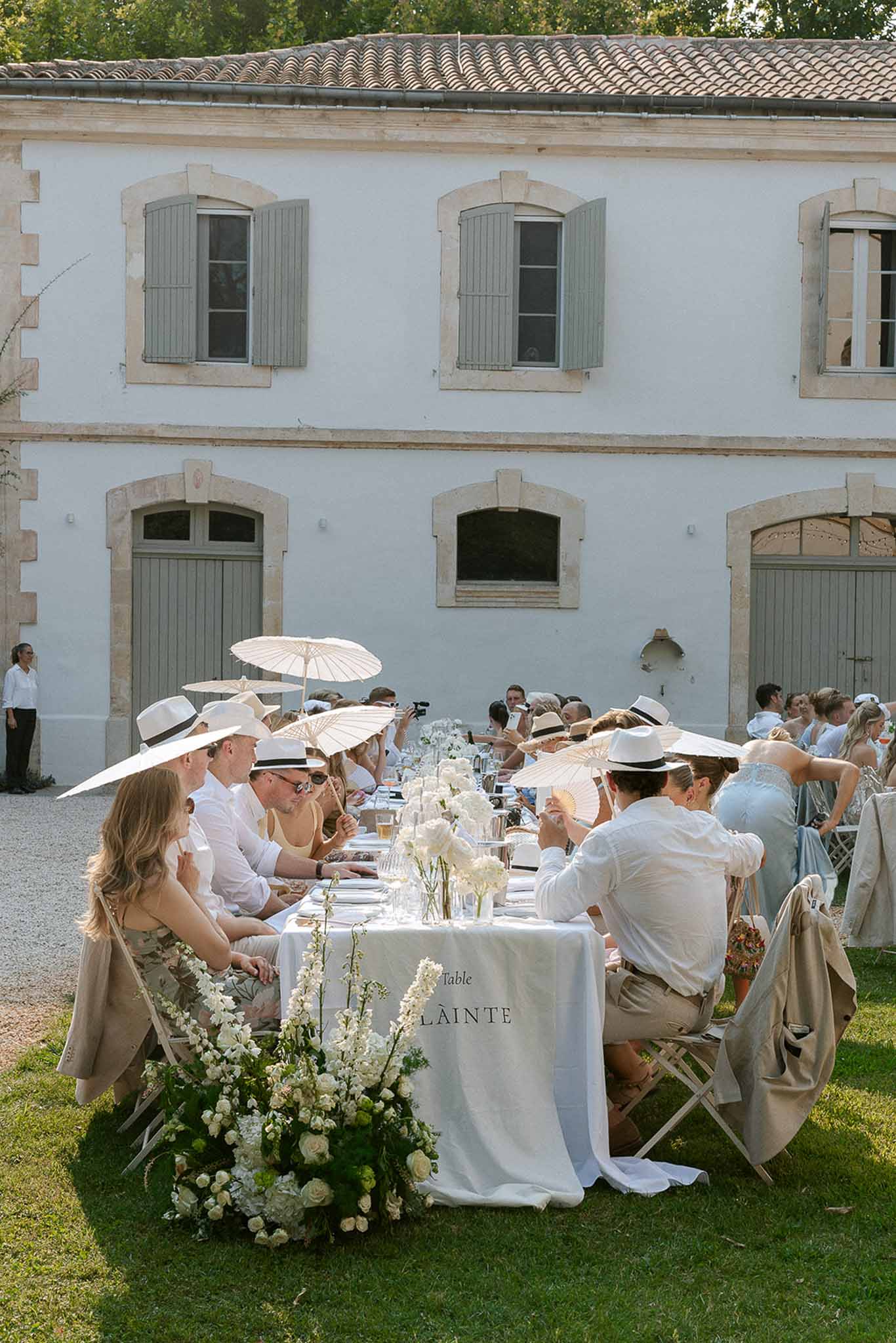 Guests in all-white attire seated at long banquet table with white roses in French estate courtyard