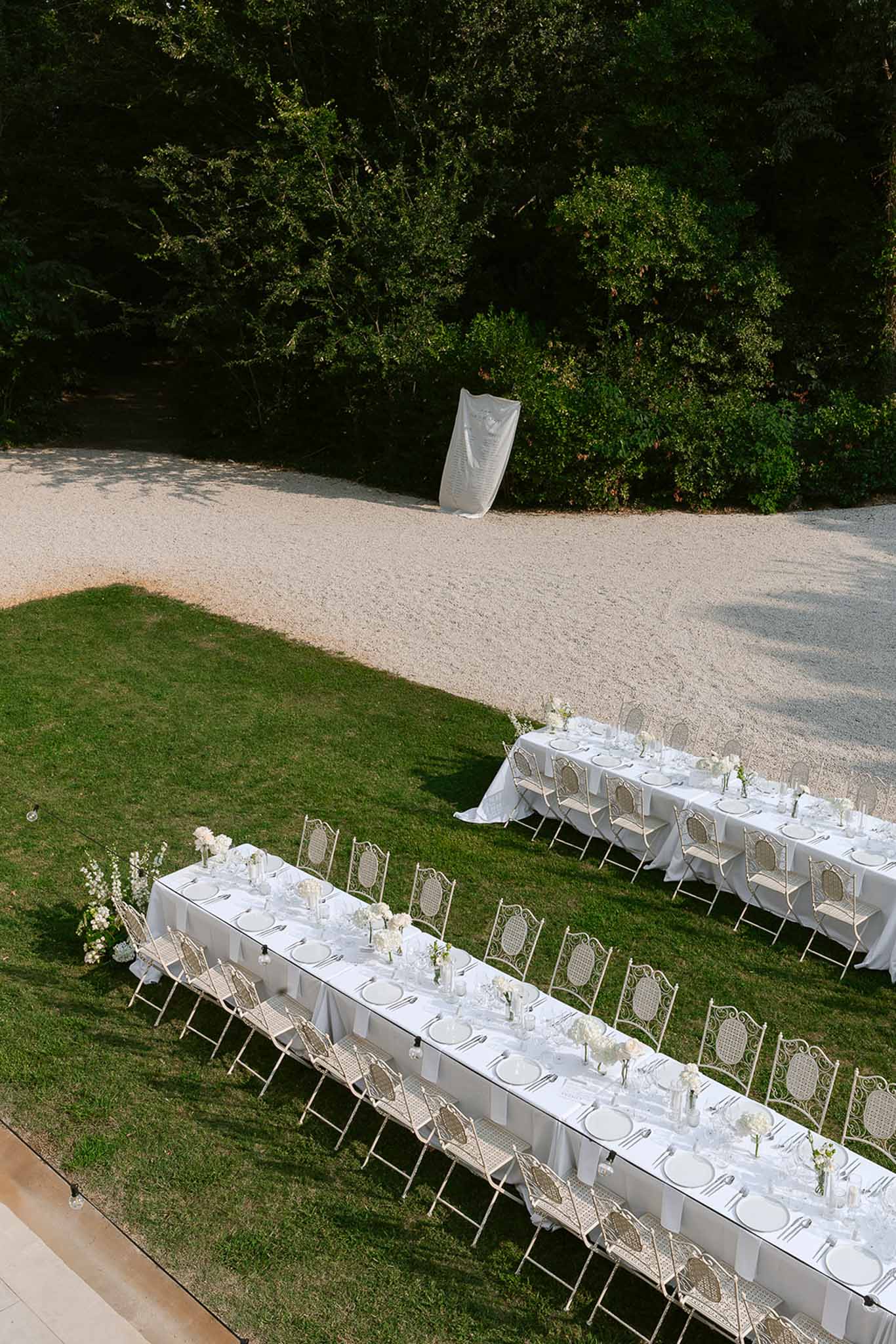 Aerial view of two long white reception tables with wrought-iron chairs and white hydrangea centerpieces on a garden lawn