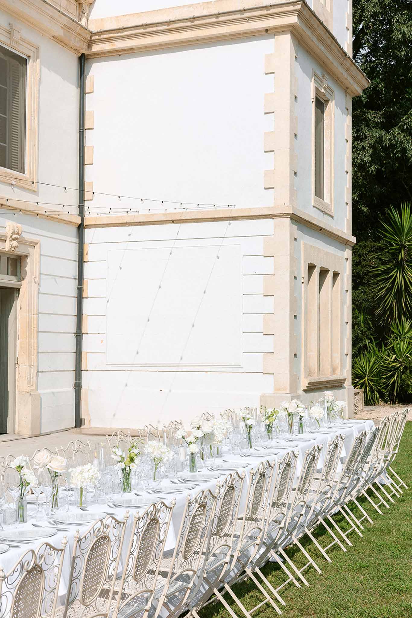 Long outdoor reception table with white roses and hydrangea centerpieces and wrought iron chairs at a French manor