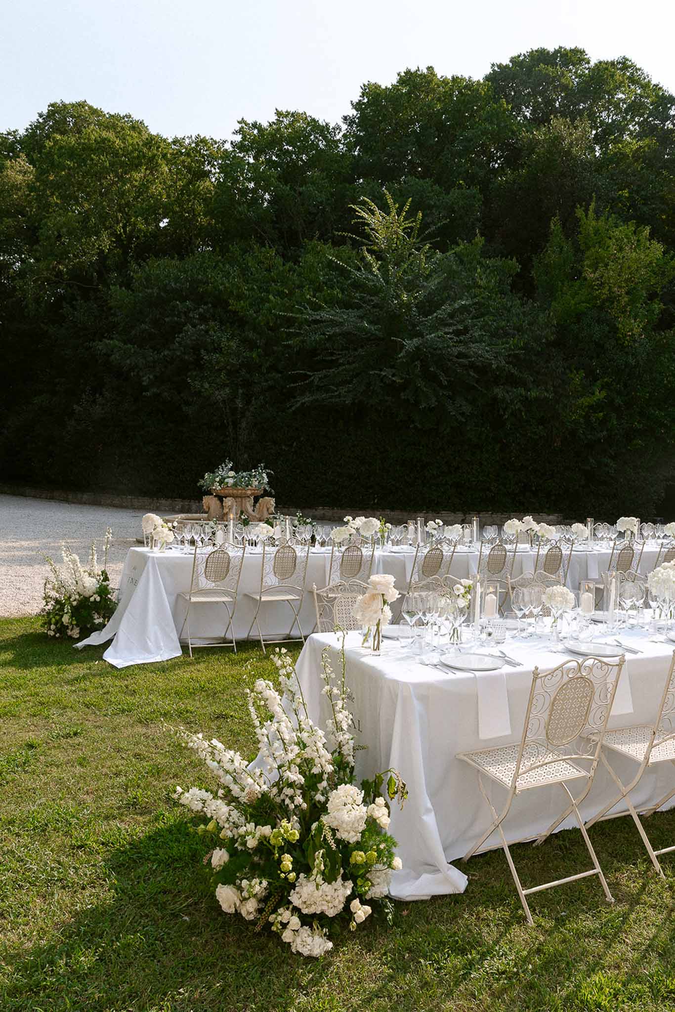 All-white garden reception with hydrangea and delphinium centerpieces cream wrought-iron chairs pillar candles on lawn