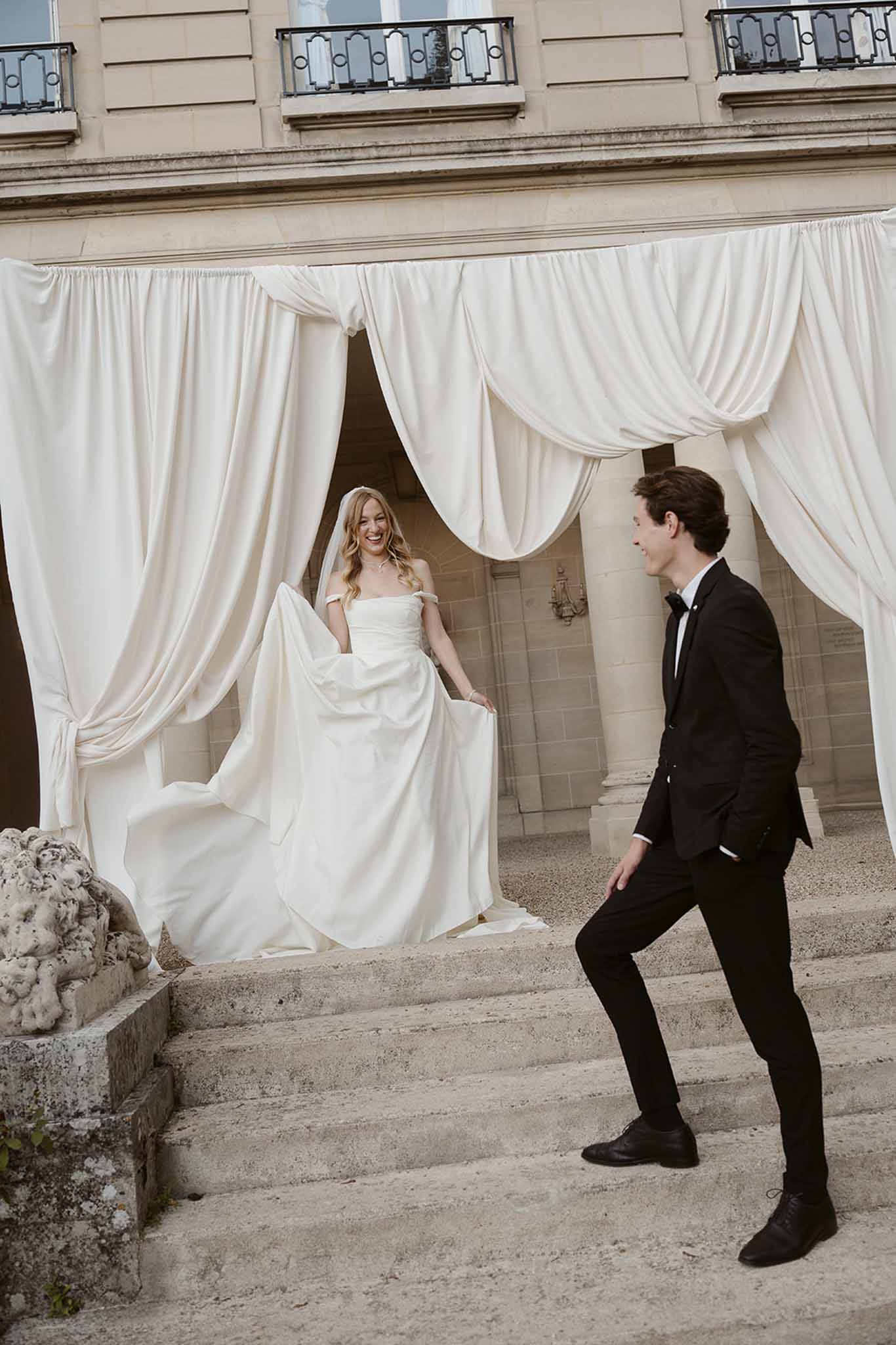 Laughing bride in off-shoulder gown on chateau steps with ivory drapes as groom climbs toward her
