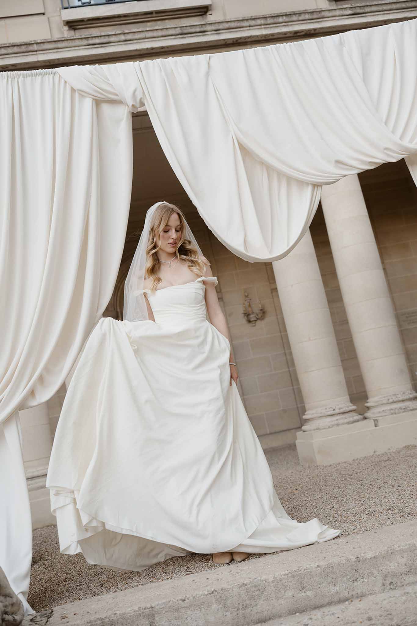 Bride in ivory off-shoulder ballgown beneath draped fabric canopy between classical stone columns