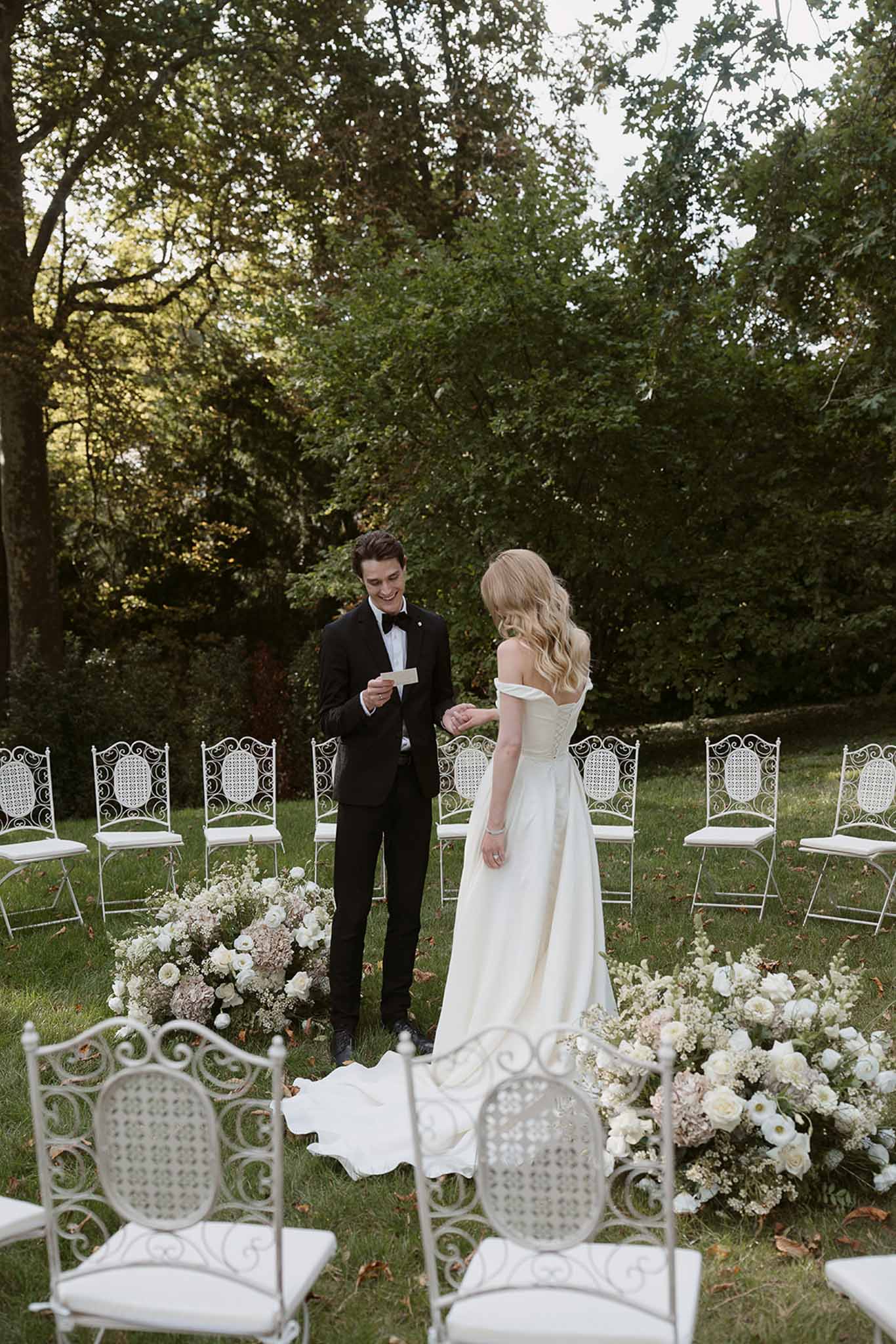 Groom reading vows holding bride's hand at garden ceremony with white ranunculus aisle florals