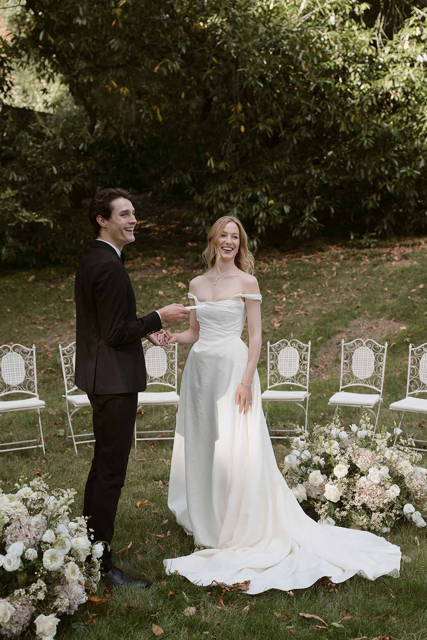 Bride and groom laughing and holding hands at outdoor garden altar lined with white roses and blush hydrangeas