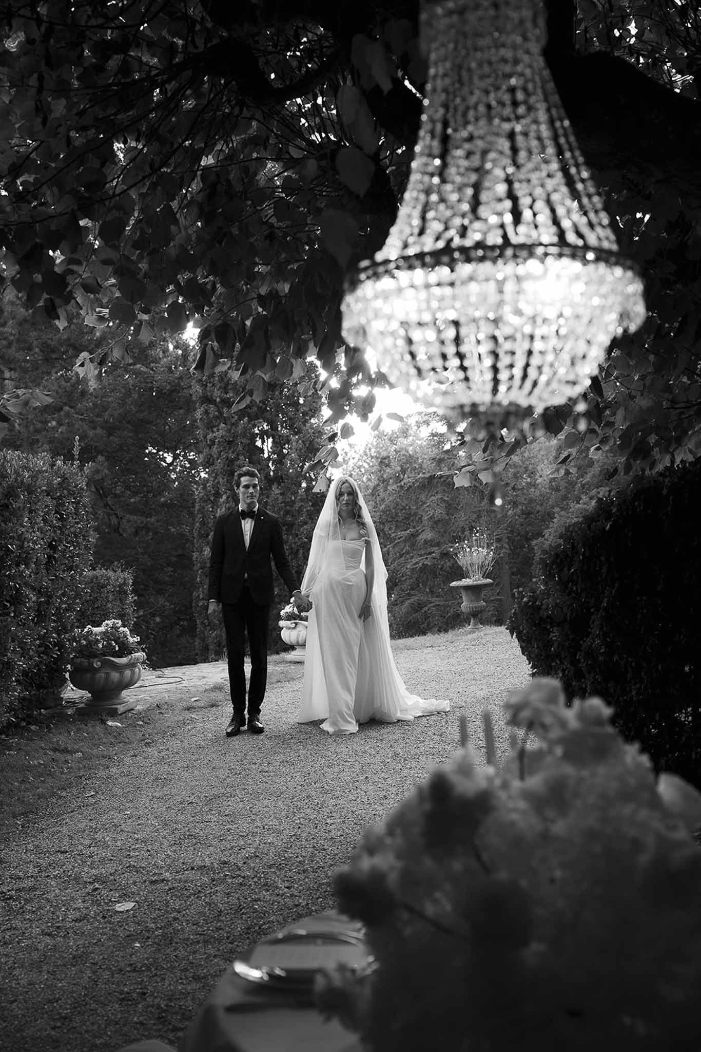 Black and white portrait of bride and groom on hedge-lined garden path with crystal chandelier hanging from tree