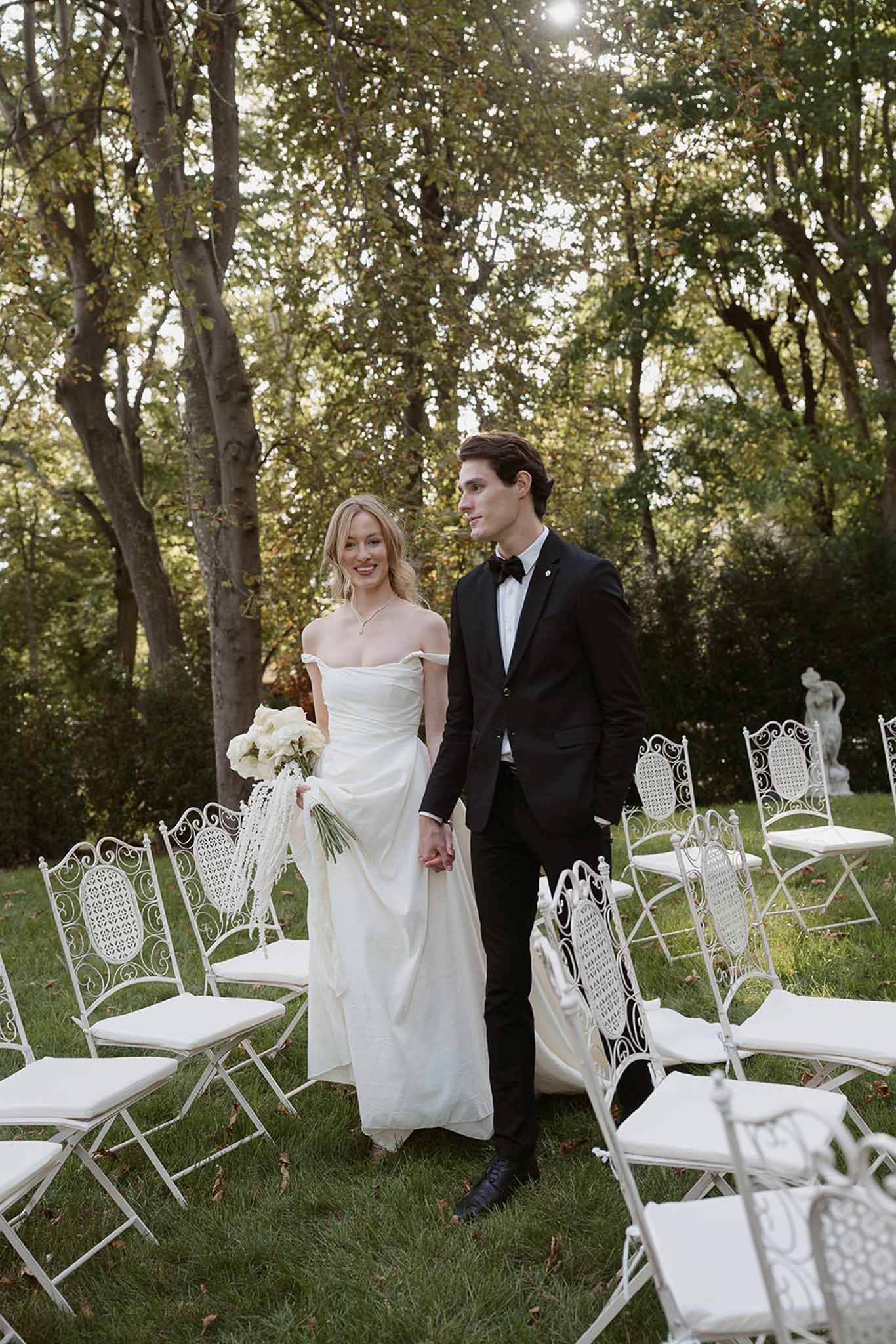 Bride in off-shoulder ivory gown with trailing ribbon bouquet and groom in black tuxedo walking down garden aisle