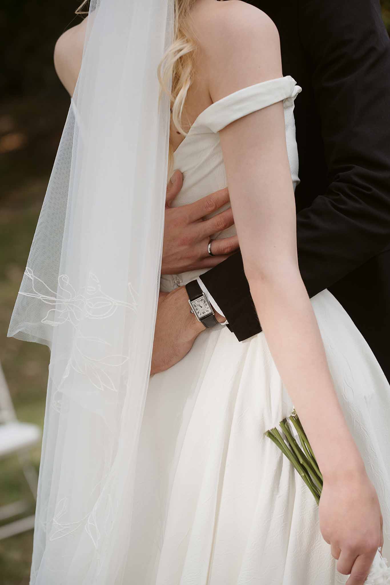 Close-up of couple embracing showing embroidered veil edge, tulip bouquet, and groom's dress watch