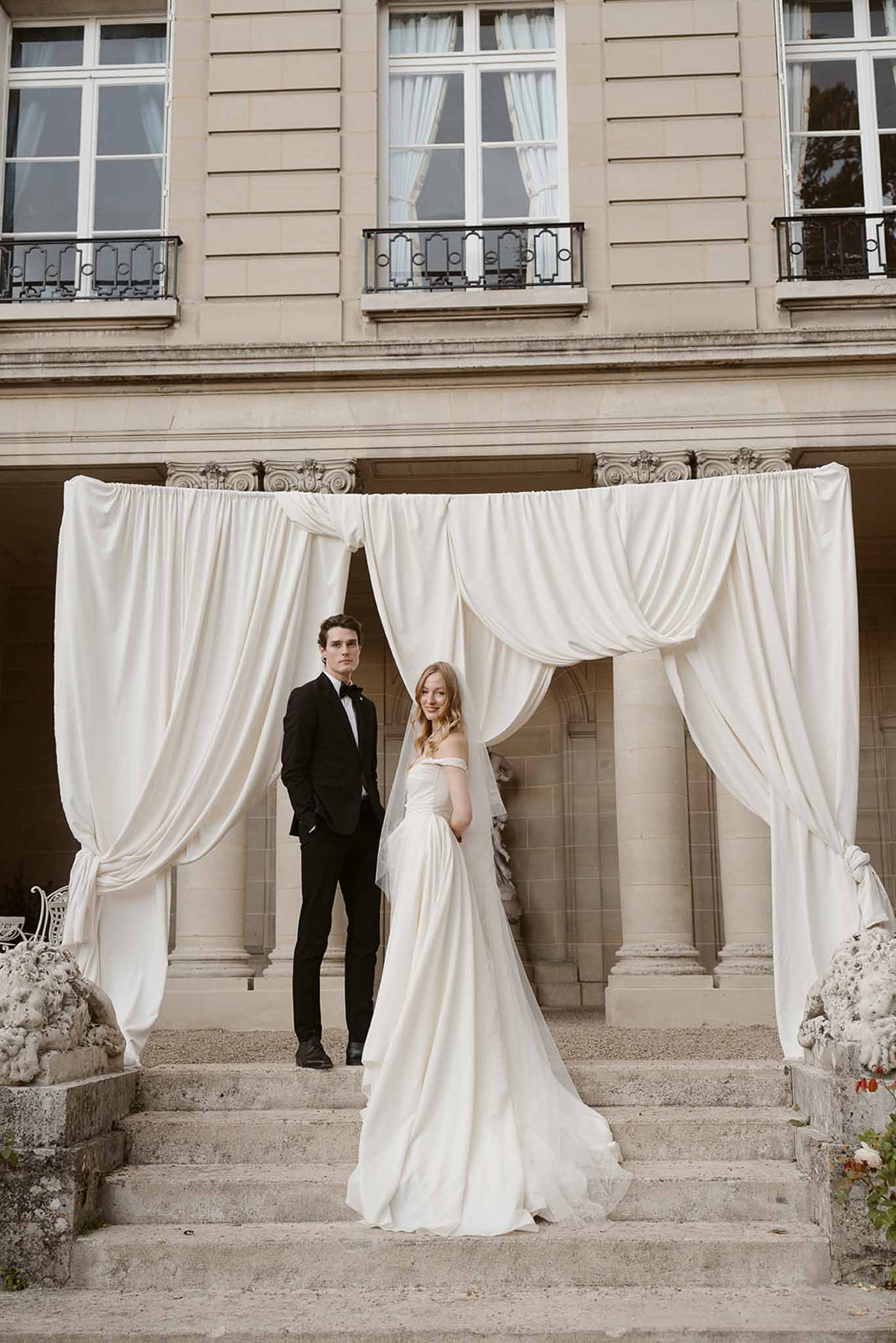 Bride and groom standing on chateau steps beneath draped ivory fabric arch with neoclassical facade behind