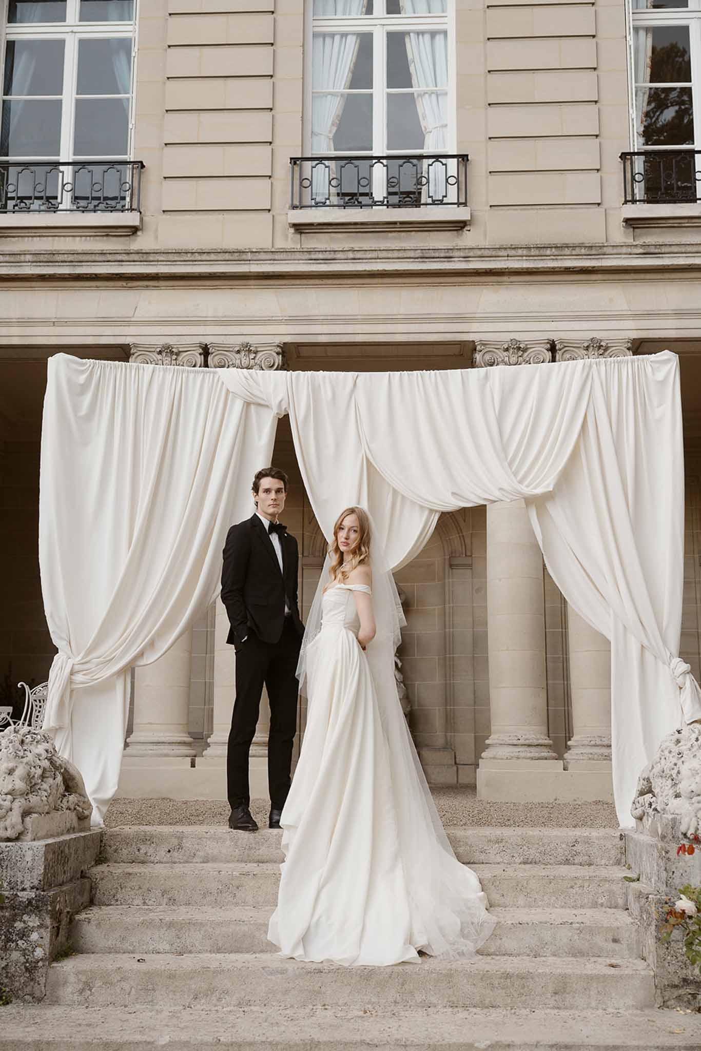 Bride and groom posing on chateau steps beneath draped ivory fabric arch, bride in off-shoulder gown with veil