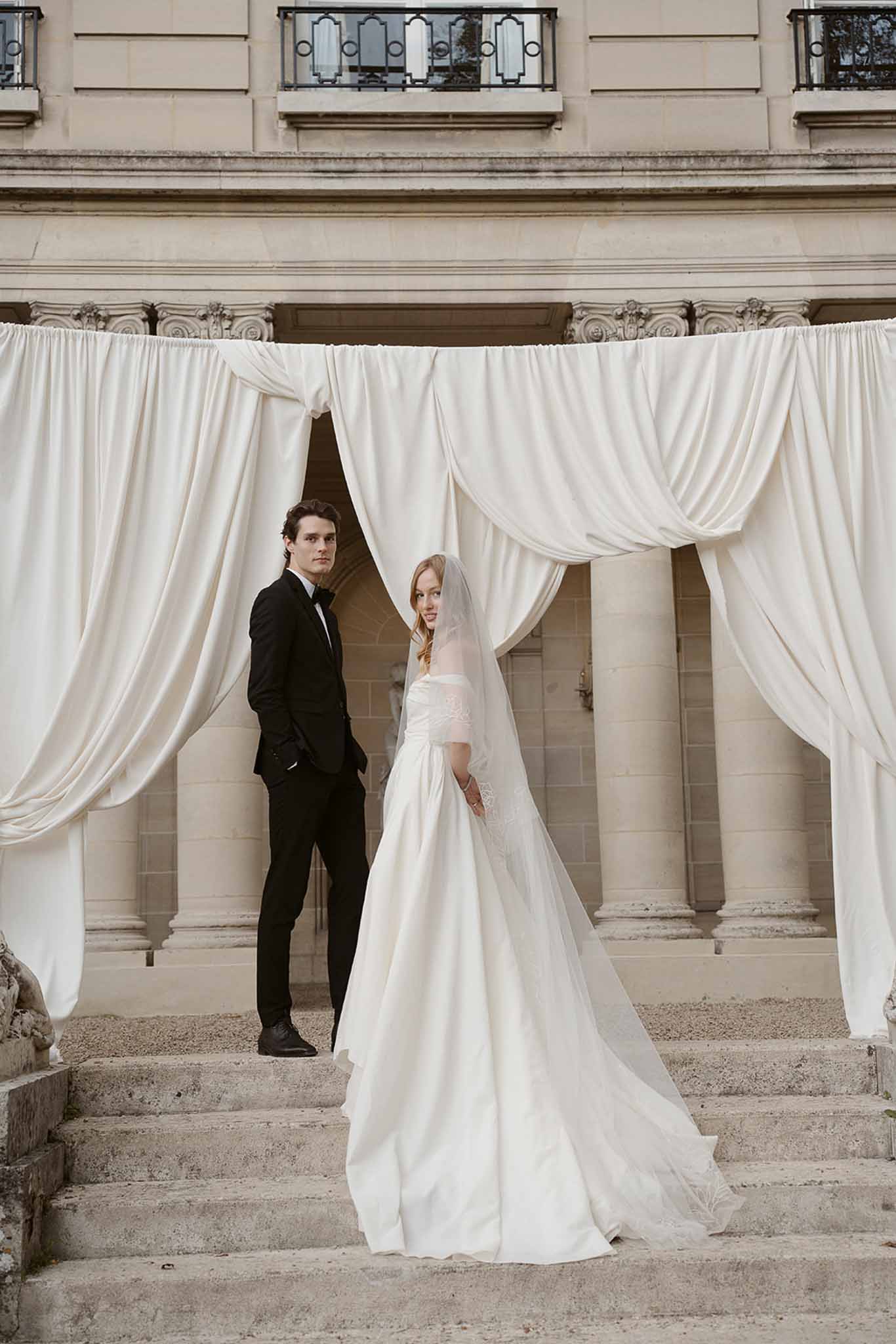 Bride in off-shoulder ivory ball gown and groom in black tuxedo posing on chateau stone steps with draped fabric