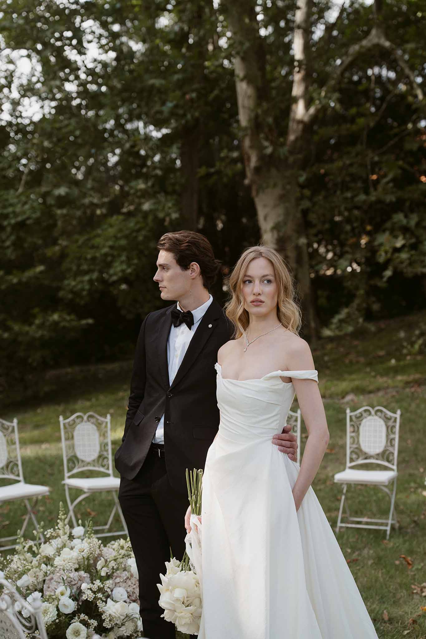 Bride with white tulip bouquet and groom in black tuxedo beside iron chairs and ranunculus arrangement