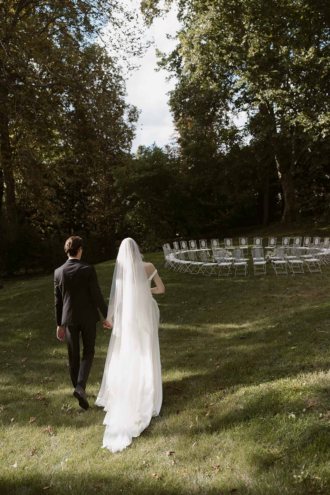 Bride and groom walking hand-in-hand toward outdoor ceremony with curved seating in a wooded garden setting
