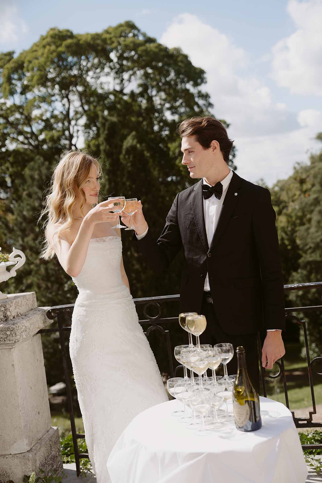 Couple toasting champagne coupes beside coupe tower on chateau terrace with wrought-iron railing