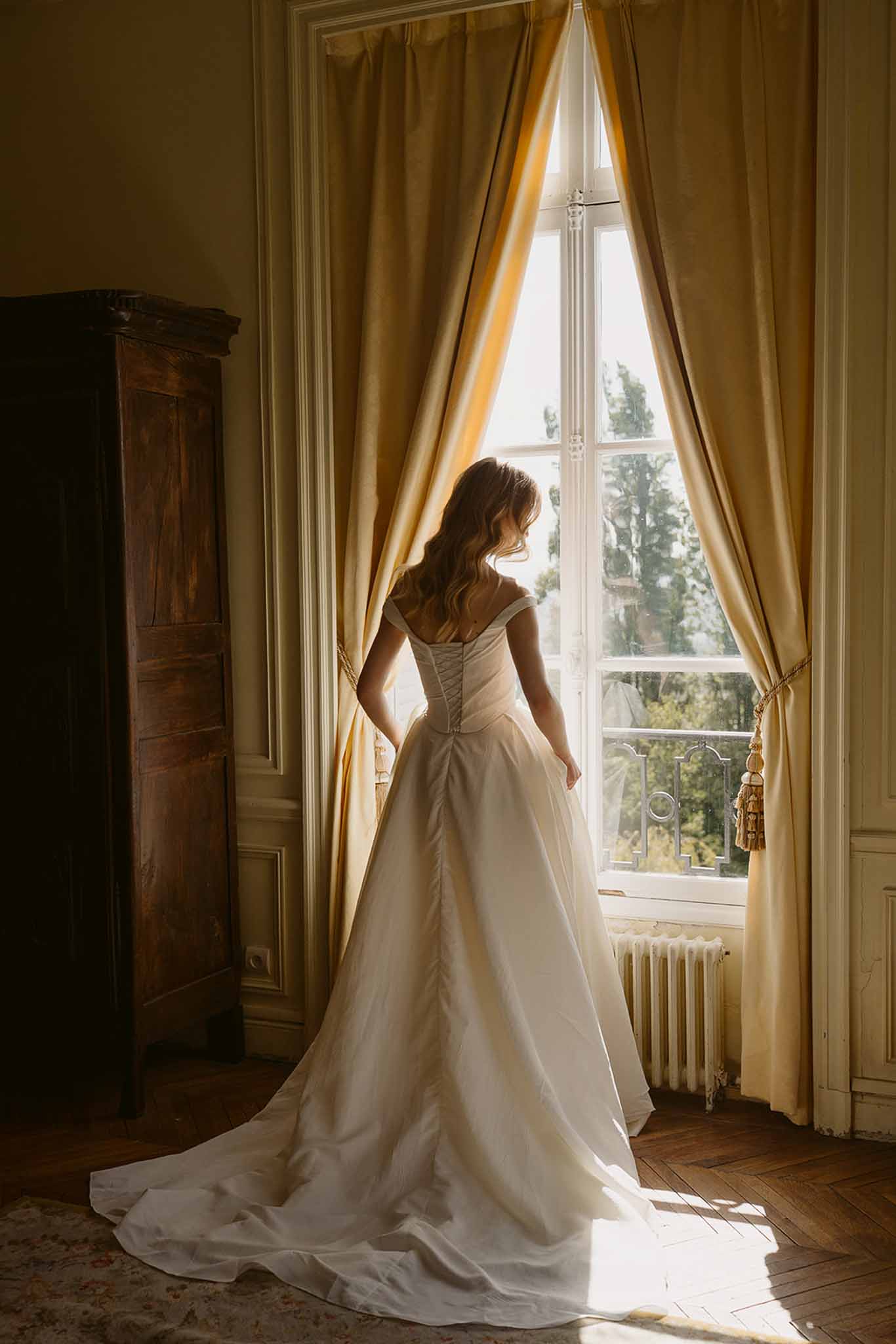 Bride in off-shoulder ball gown with corset back and cathedral train looking out tall window in French chateau room