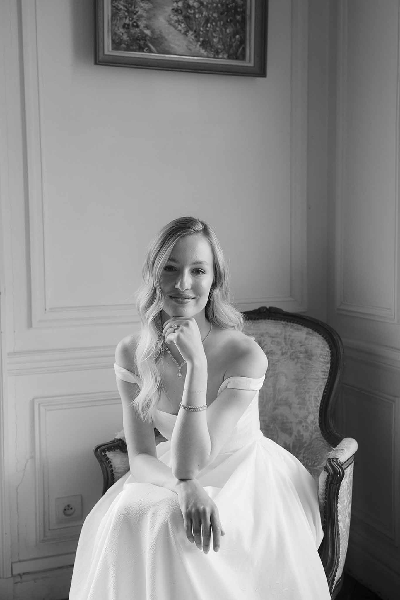 Bride in off-shoulder ballgown seated in Louis XVI armchair with panelled walls and painting in B&W