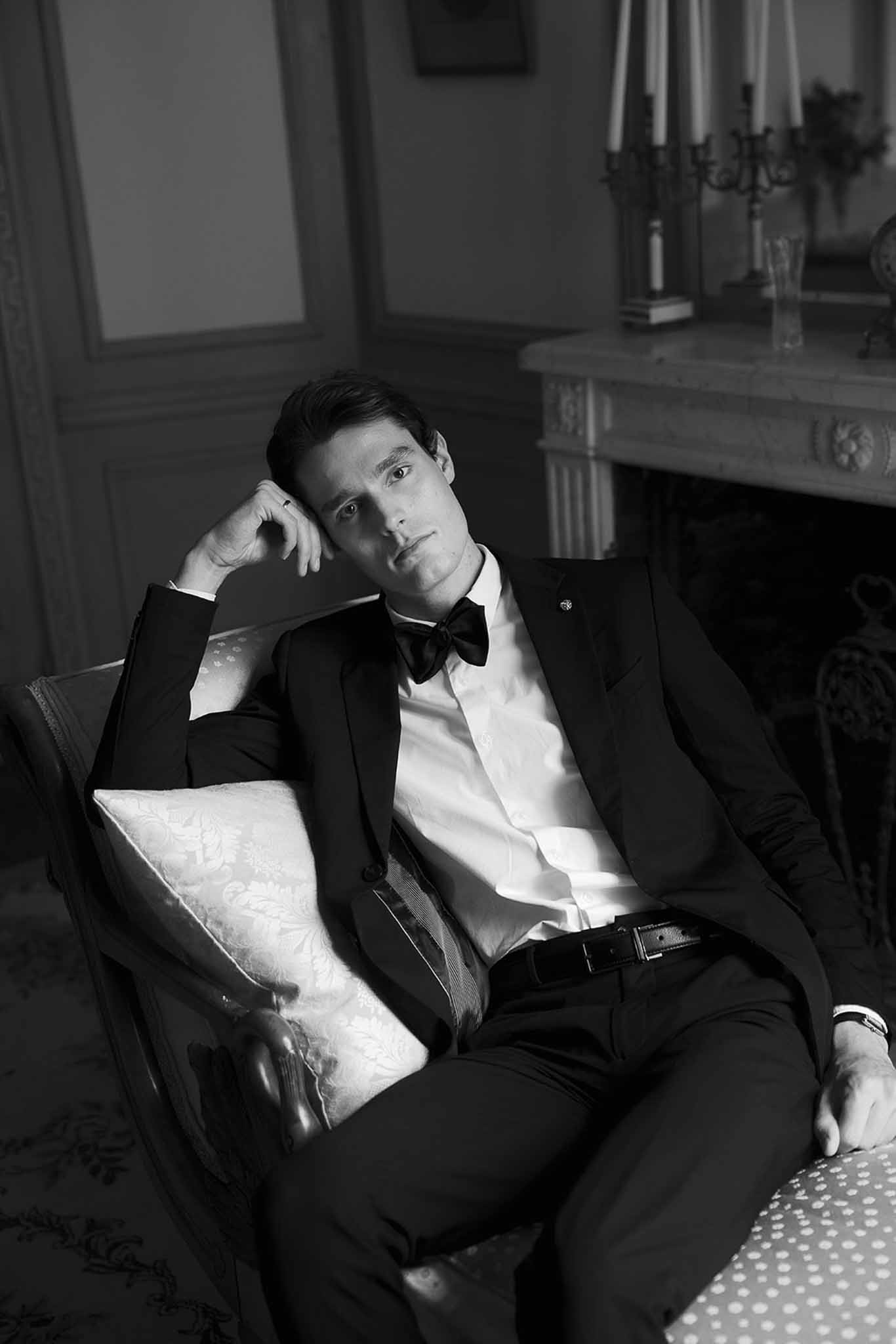 Black and white portrait of groom in tuxedo seated in armchair beside candelabra on fireplace