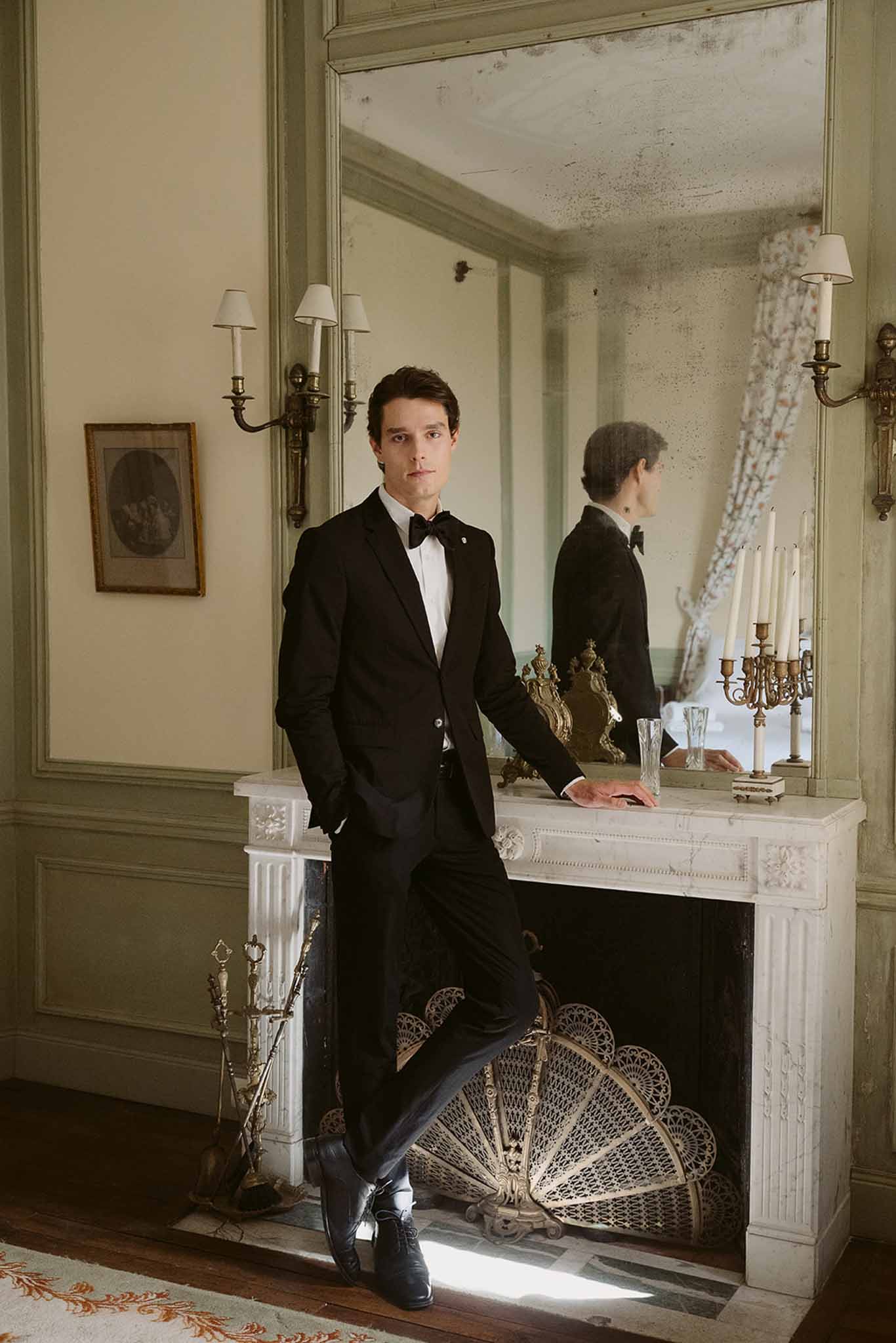 Groom in black tuxedo leans against marble fireplace in sage-paneled room with antique mirror and clock