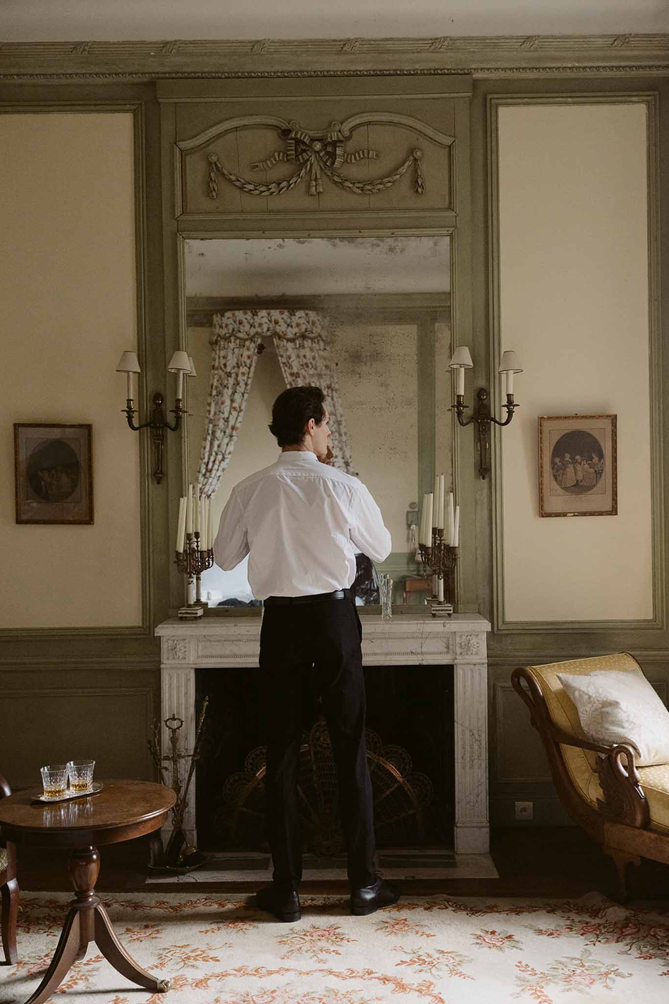Groom from behind at marble fireplace adjusting cuffs in room with sage boiserie foxed mirror and brass sconces