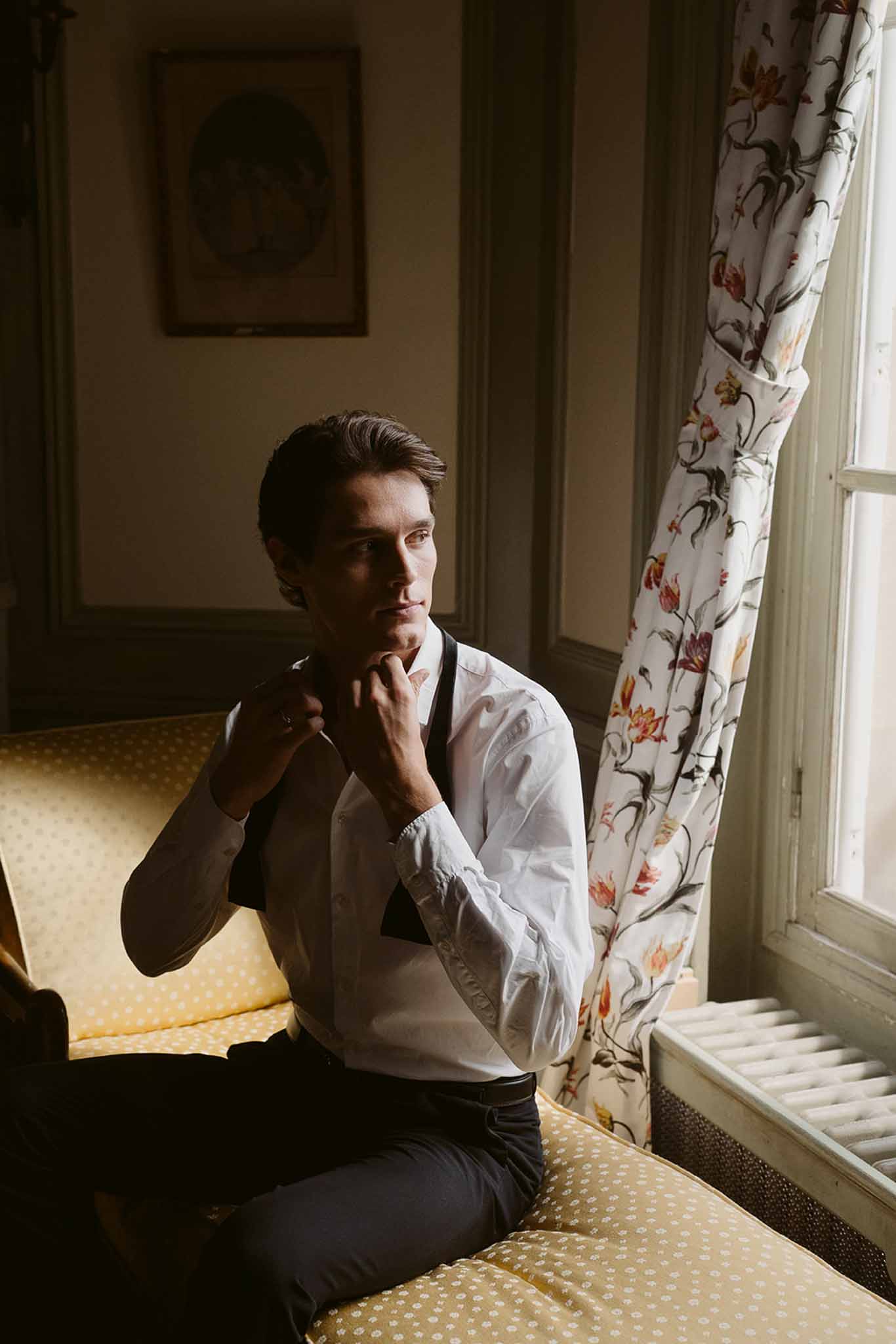 Groom in white shirt and suspenders adjusting collar by window in room with tulip-print curtains