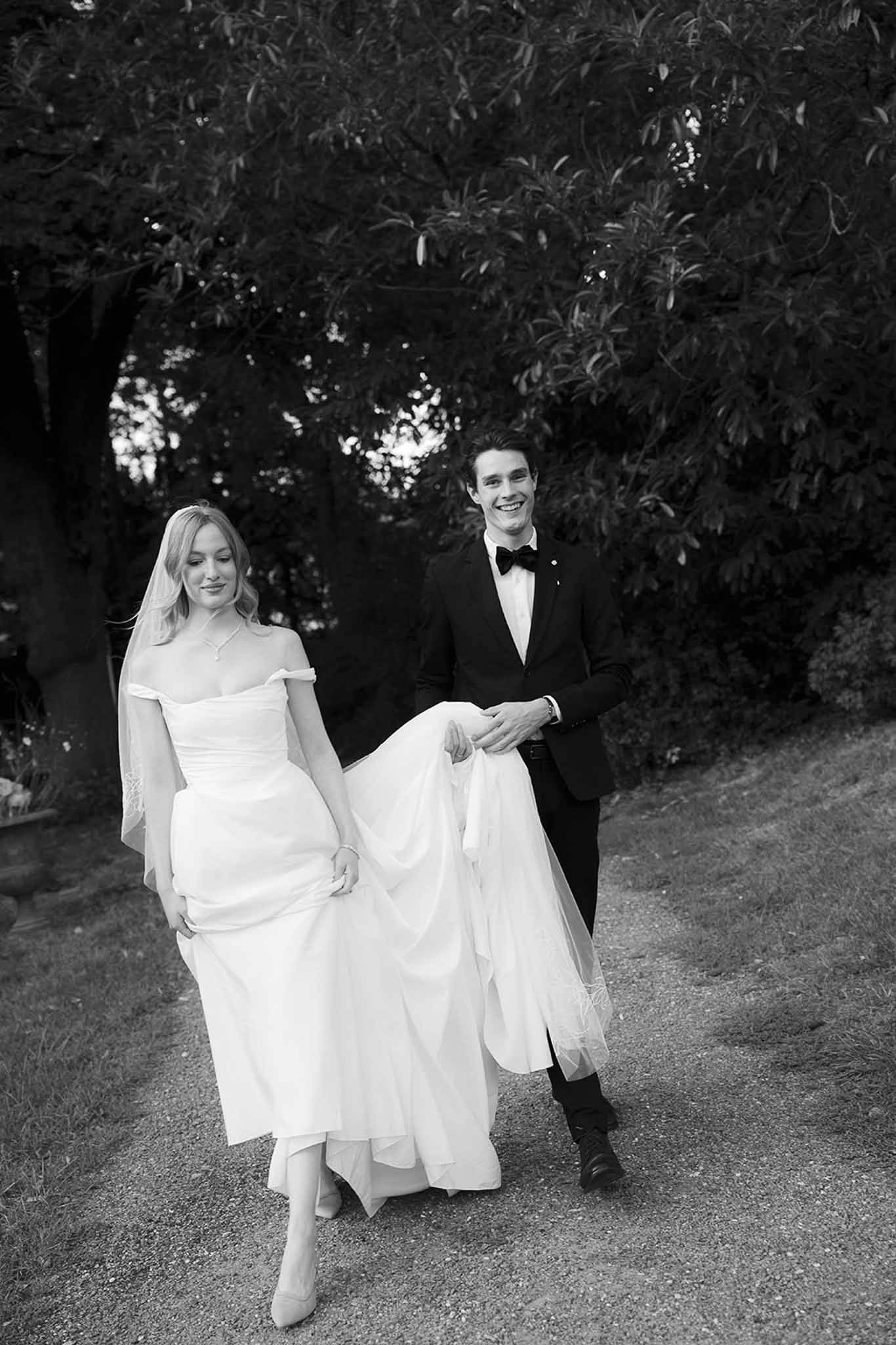 Black and white portrait of groom carrying bride's train as they walk along tree-lined gravel path
