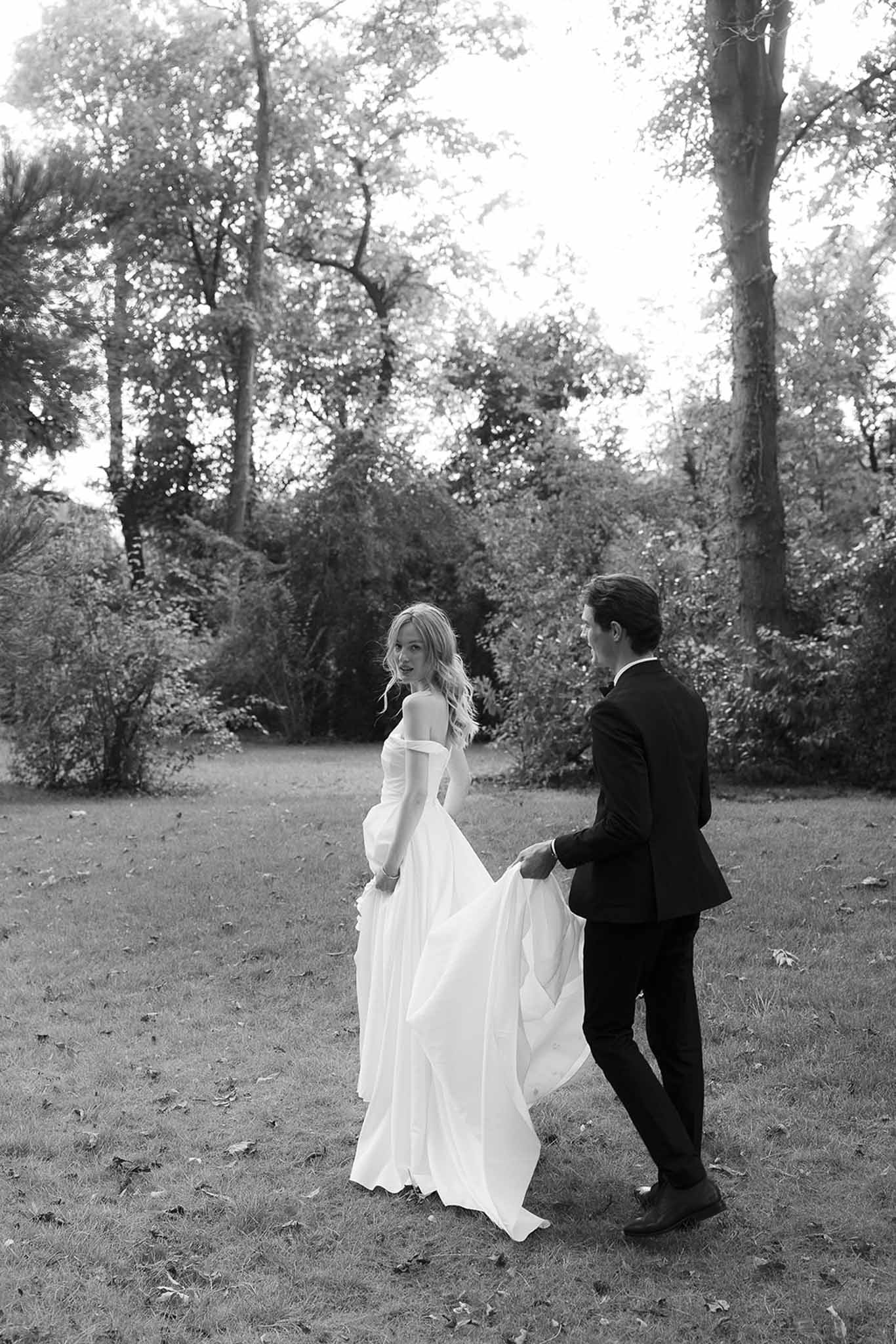Black and white photo of bride and groom walking across garden lawn with groom holding dress train