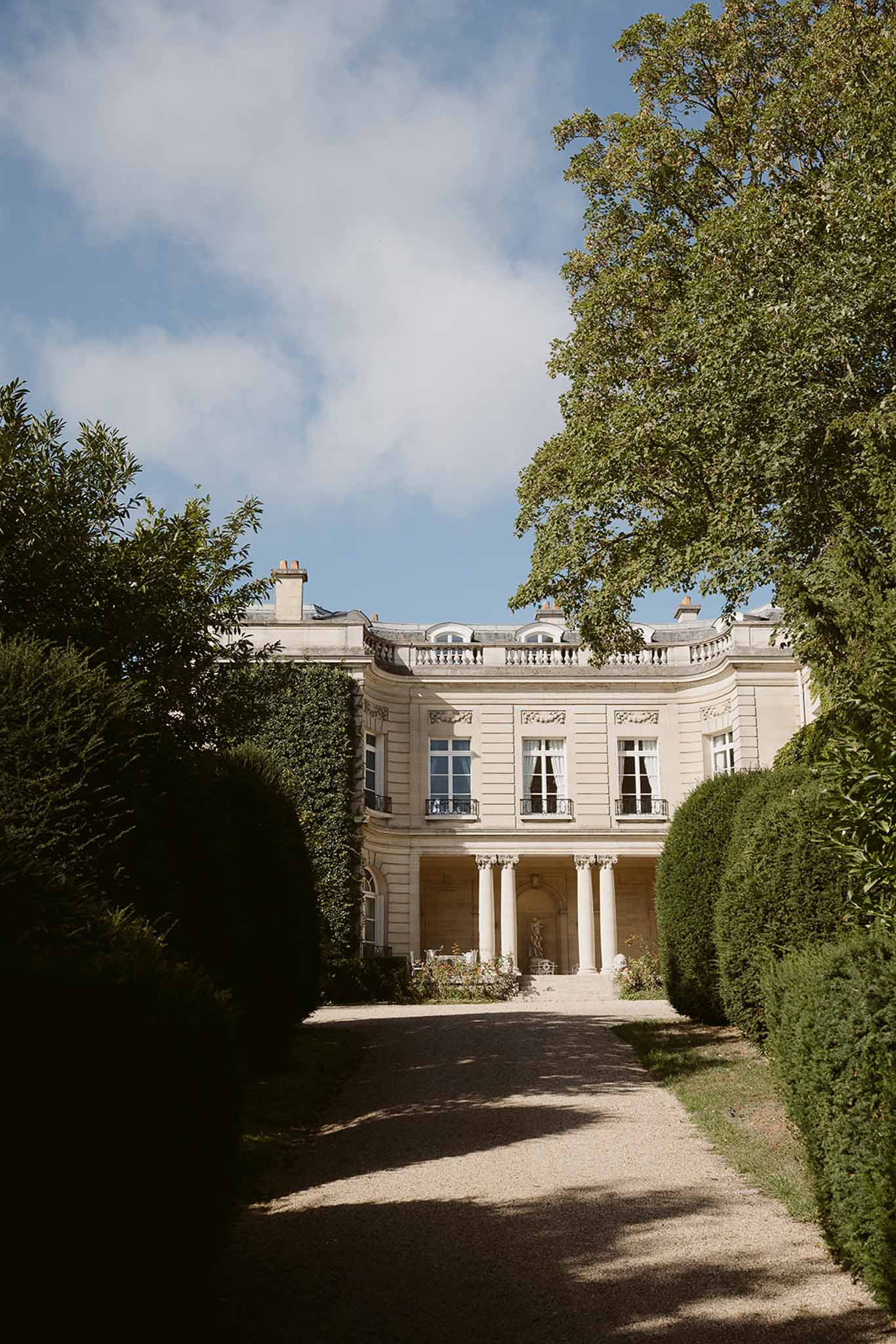 Neoclassical chateau with curved columned portico, carved friezes, and clipped hedge-lined driveway