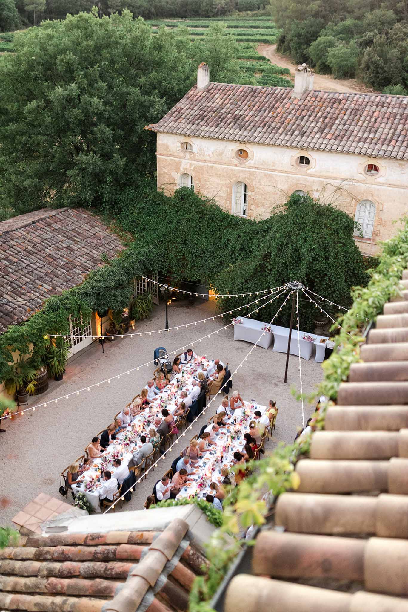 Aerial view of courtyard reception with guests at long banquet tables, festoon lights, and vineyard rows beyond