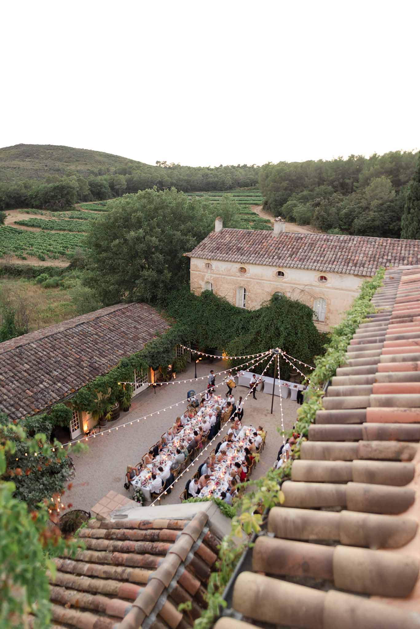 Aerial view of outdoor wedding reception dinner in a Provencal courtyard with festoon lights and vineyard backdrop