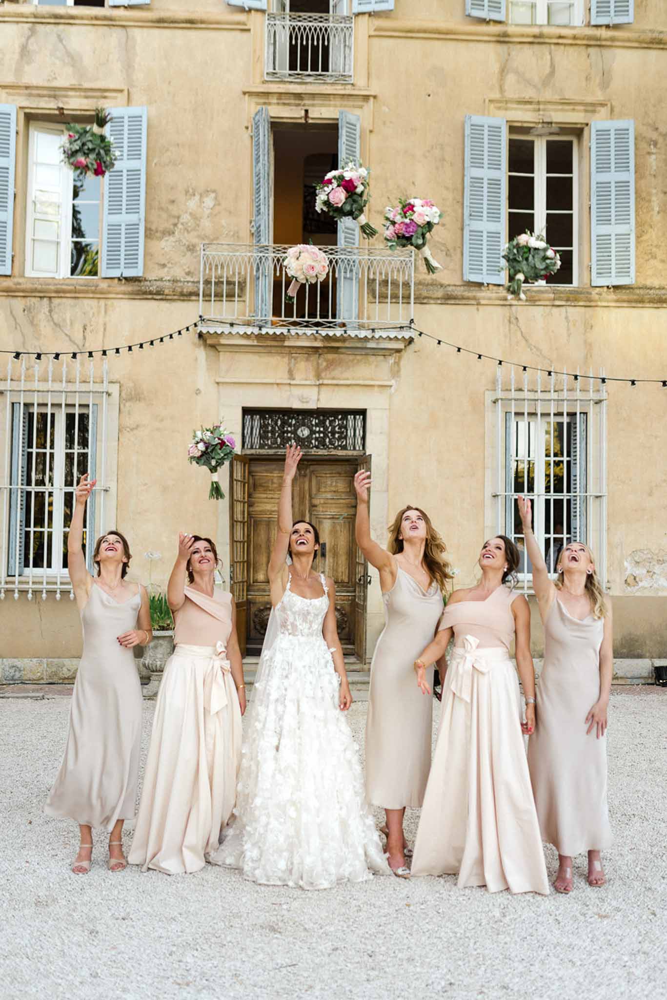 Bride and five bridesmaids tossing bouquets in air before chateau facade with festoon lights