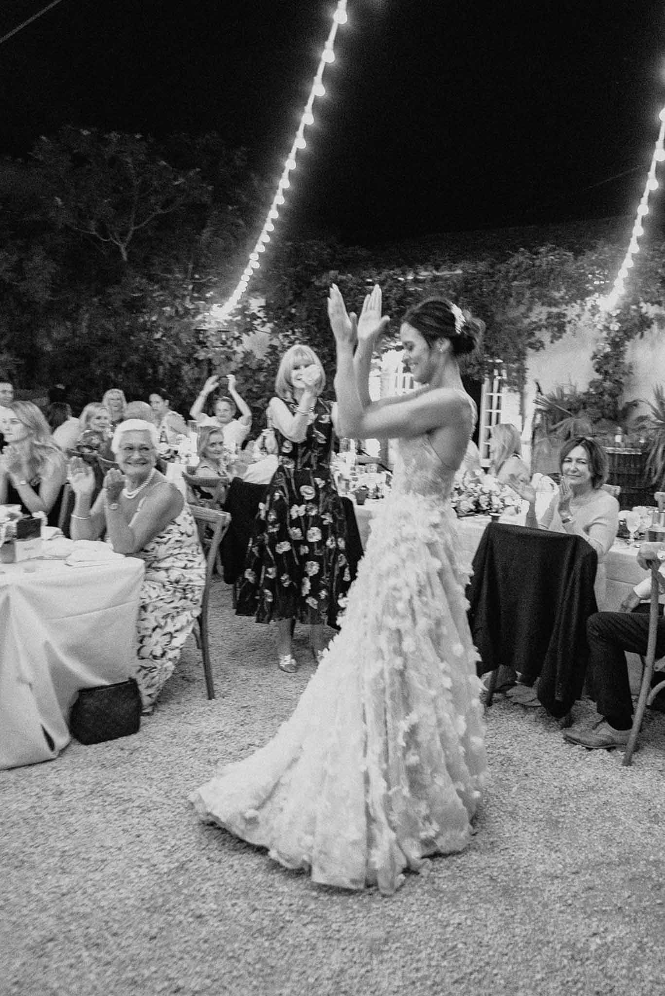 Black and white shot of bride dancing with arms raised under fairy lights at courtyard reception